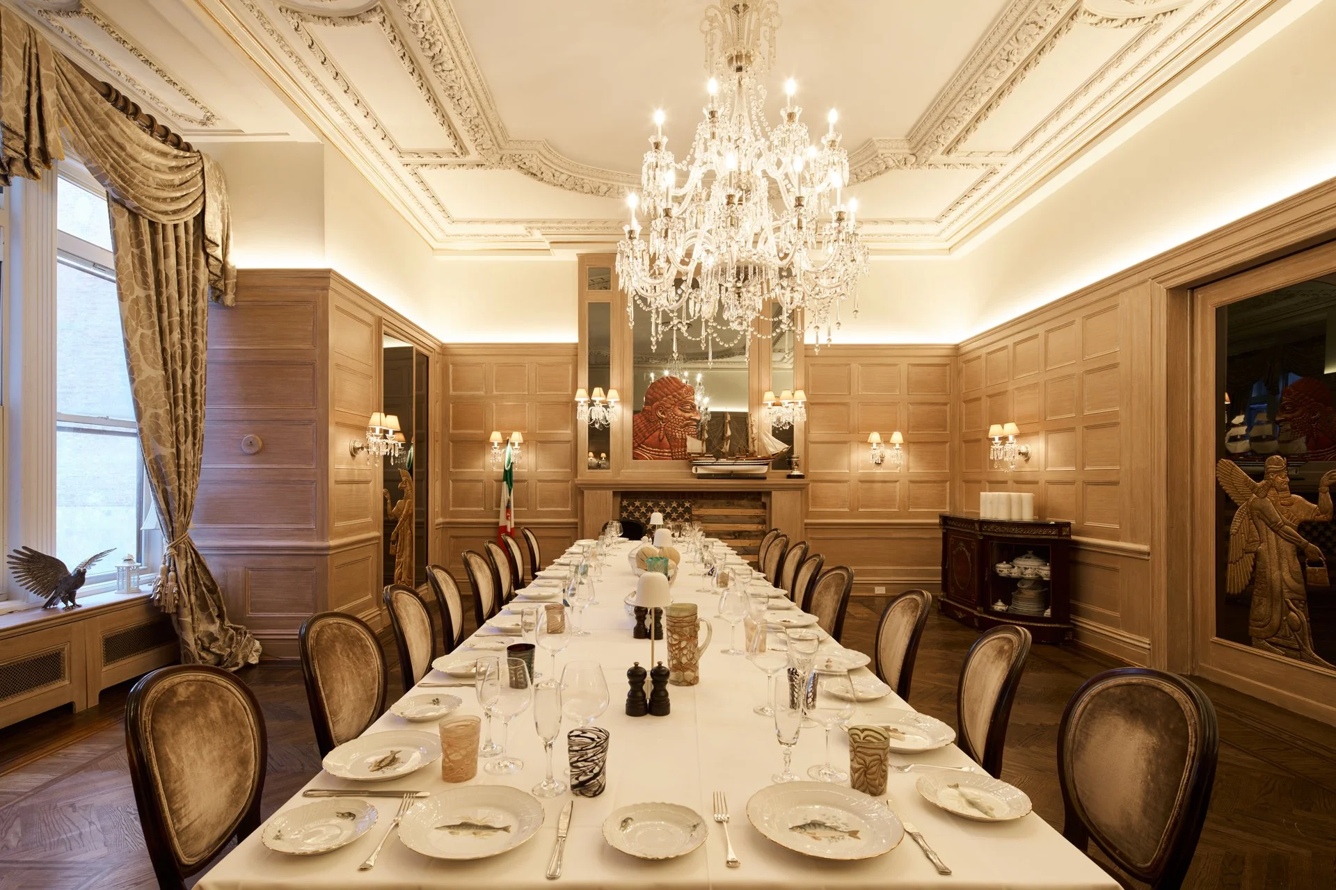 Elegant dining room with a long table set for a meal, ornate ceiling moldings, a large chandelier, wood-paneled walls, and historical artwork and artifacts.
