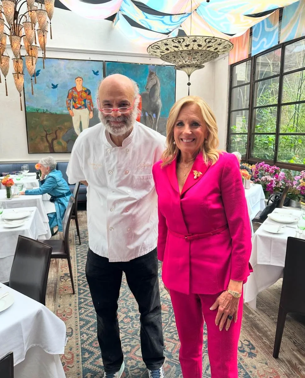A man wearing a white chef's coat and glasses, smiling, standing next to a woman in a bright pink suit with a gold pin, smiling, in a colorful restaurant with paintings, umbrellas, and flowers.
