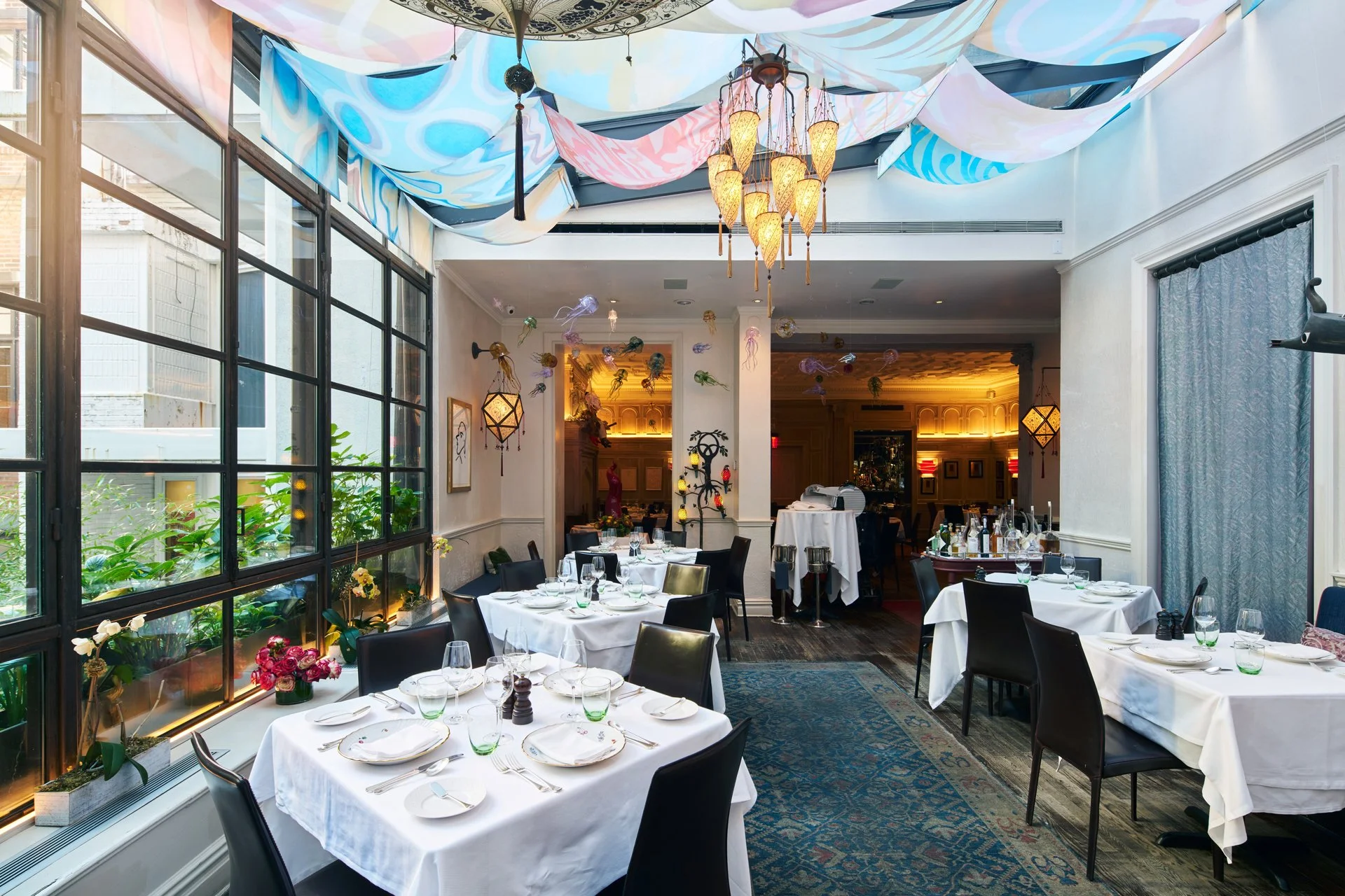 Elegant restaurant with white tablecloths, black chairs, and large windows with greenery outside, decorated with hanging lights and ceiling fabric for a festive atmosphere.