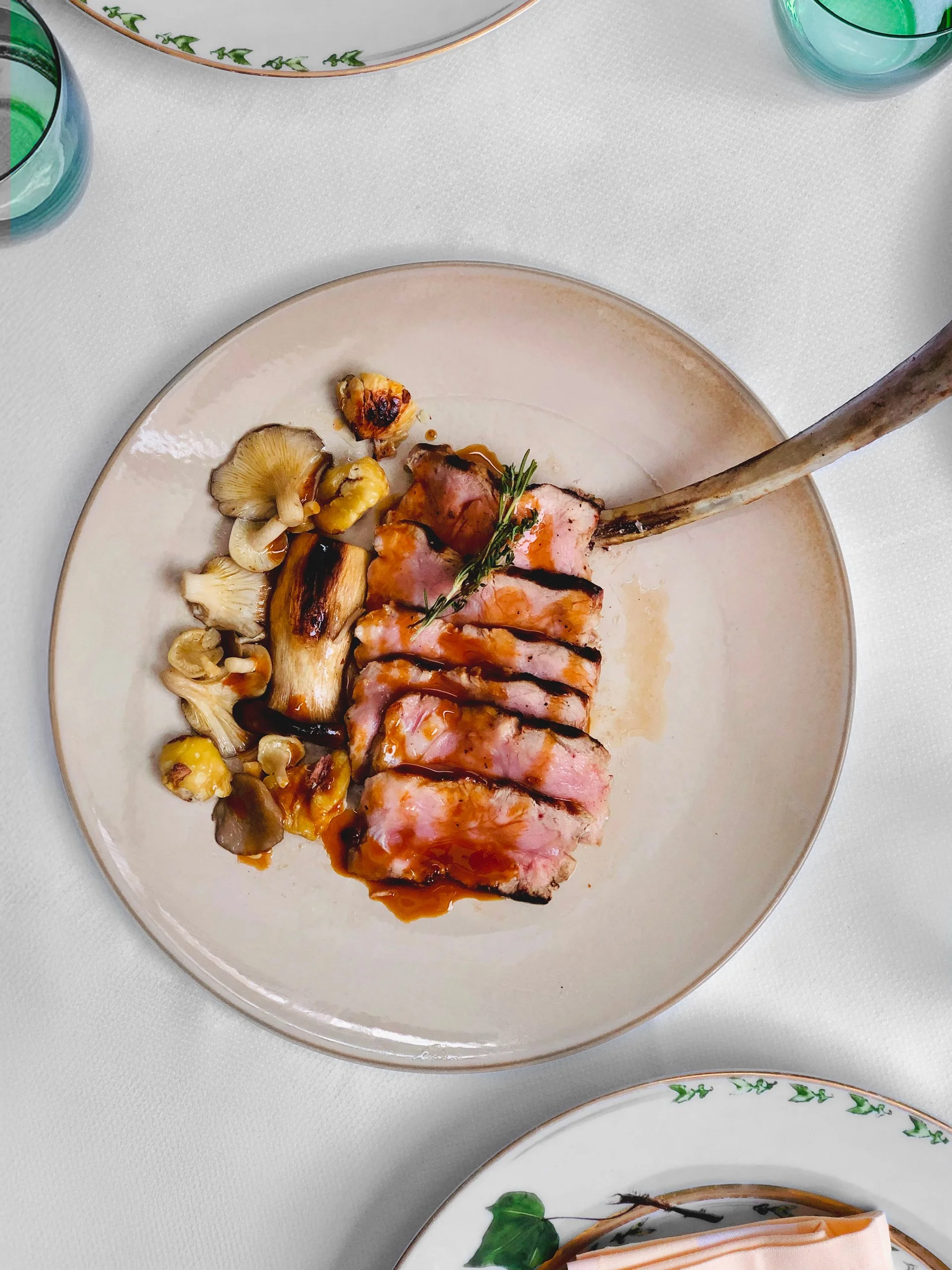 A plated dish featuring sliced, grilled rack of lamb with a sprig of rosemary garnish, served with a side of lightly sautéed wild mushrooms.