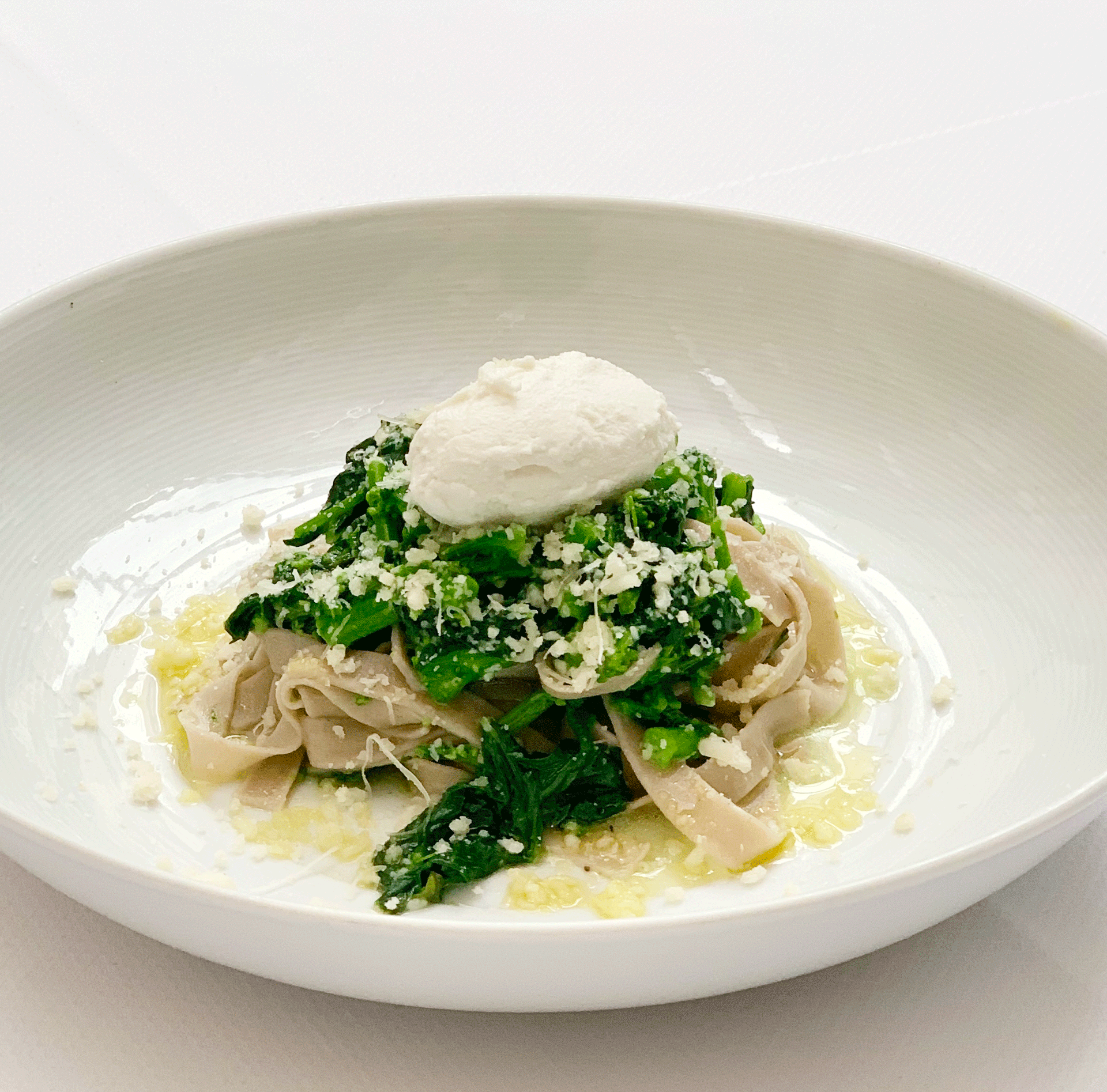 A white bowl with a gourmet pasta dish topped with green vegetables, grated cheese, and a scoop of white cheese or cream.