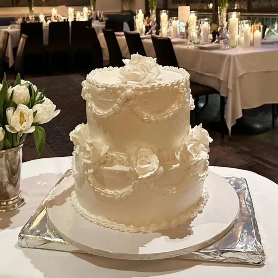 A two-tiered white wedding cake with decorative piping and floral accents, placed on a silver cake board on a white table, with a floral arrangement nearby in a silver vase and a decorated banquet hall in the background.