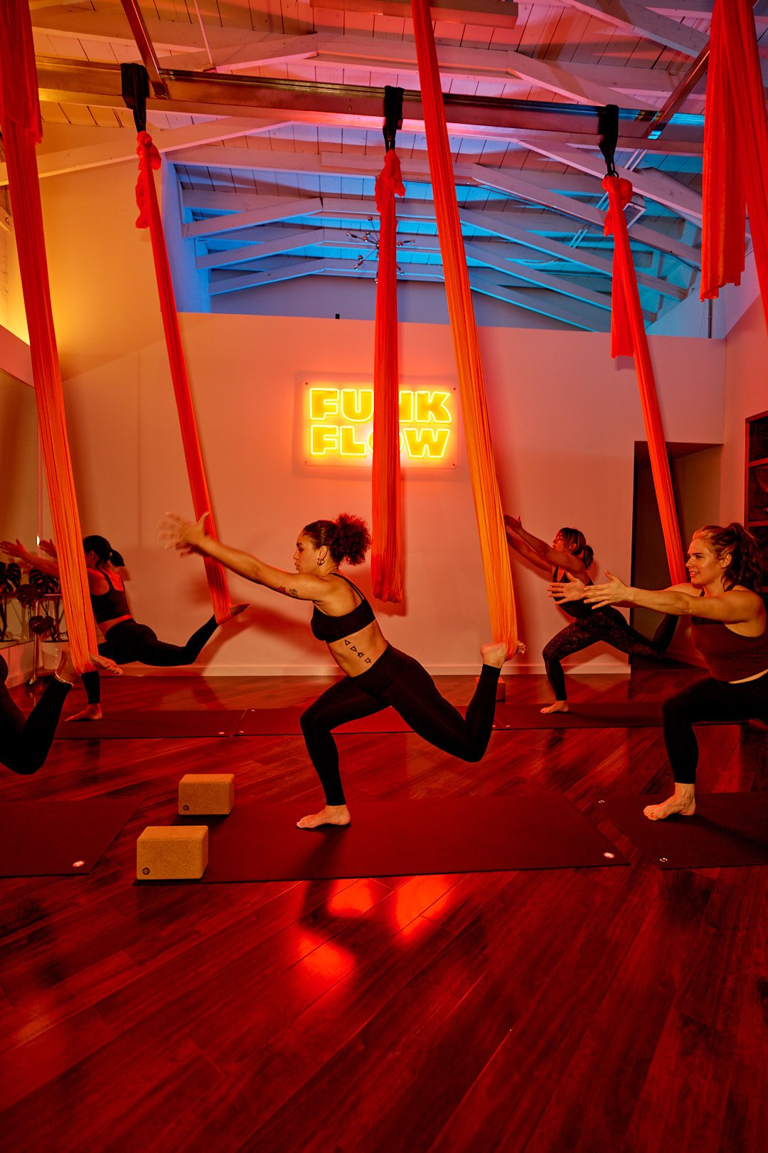 Women practicing aerial yoga with red fabric swings in a studio with neon sign that says 'FUNK FLOW' and hardwood floors.