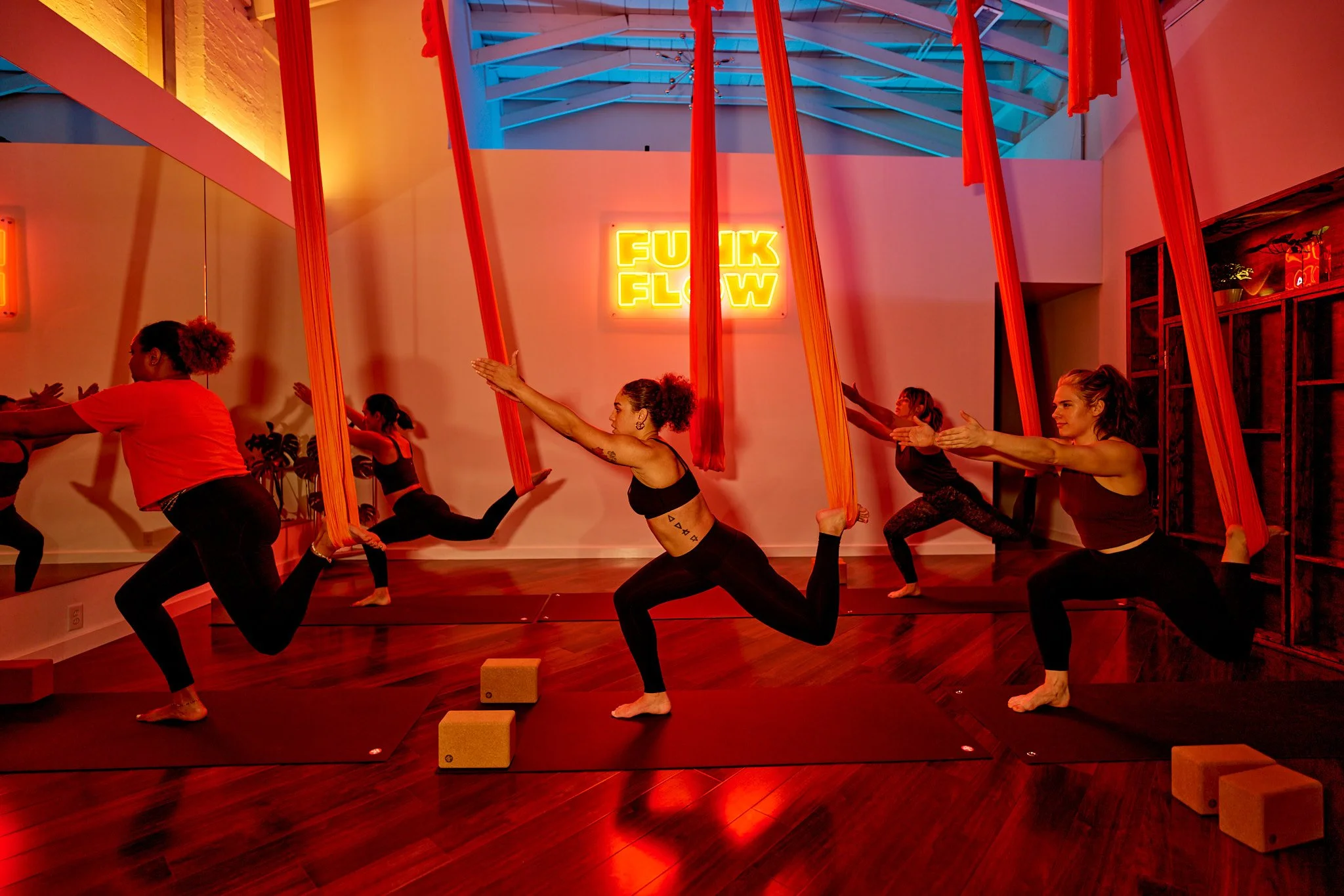 Group of women practicing aerial yoga with red fabric hammocks in a studio with a neon sign that says 'Funk Flow' in the background.