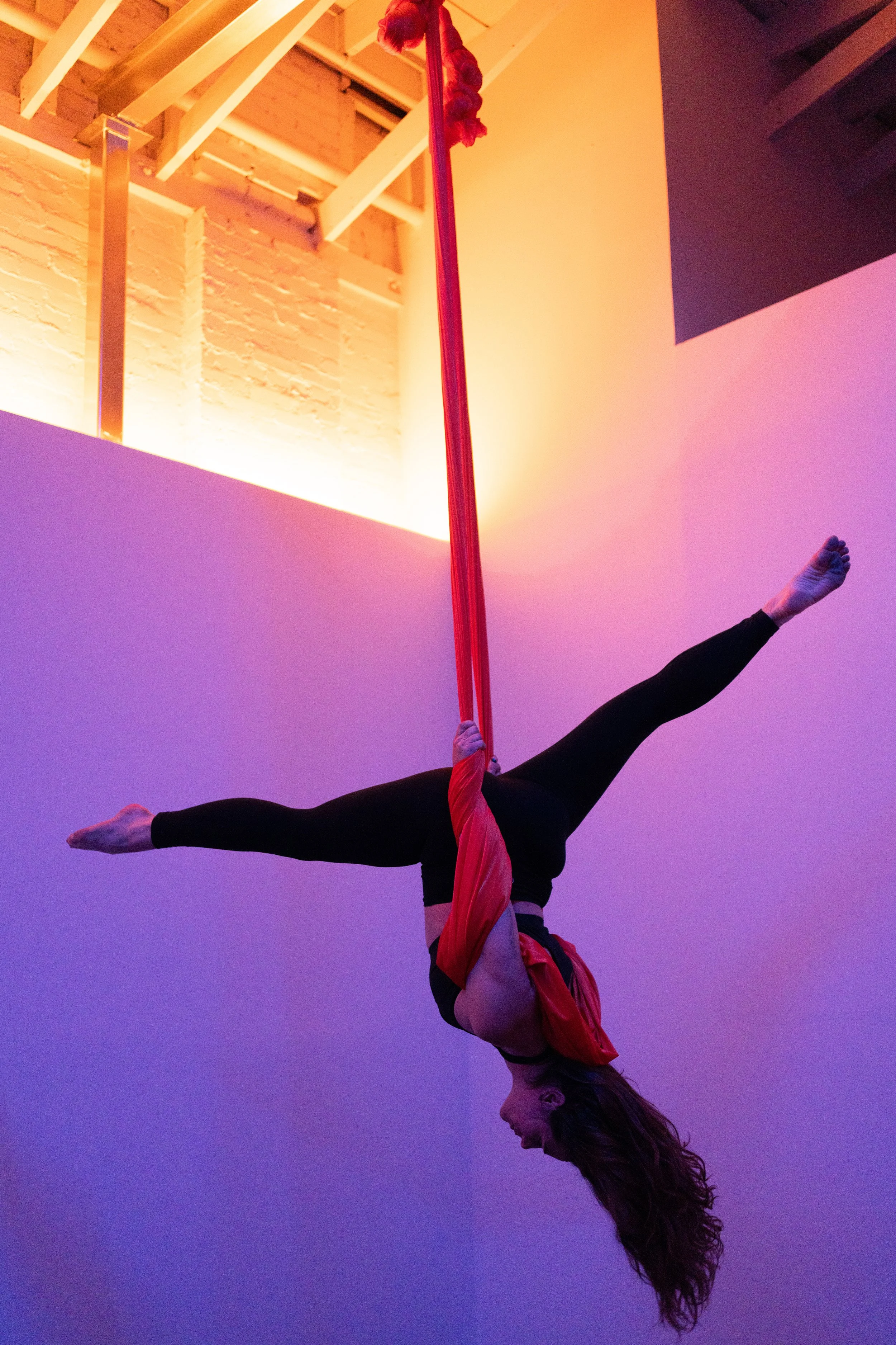 A woman performs an aerial silk act on red silks hanging from the ceiling in a colorful, dimly lit room.