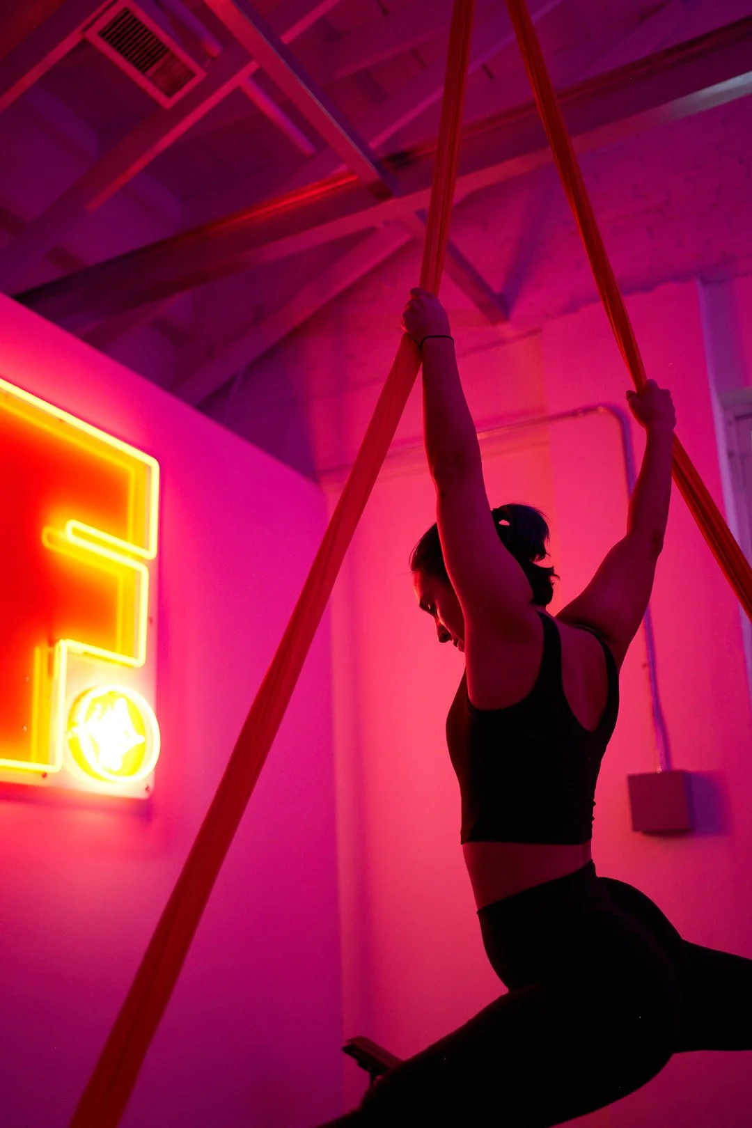 A woman practicing aerial silks in a studio with pink and purple lighting and a neon sign on the wall.
