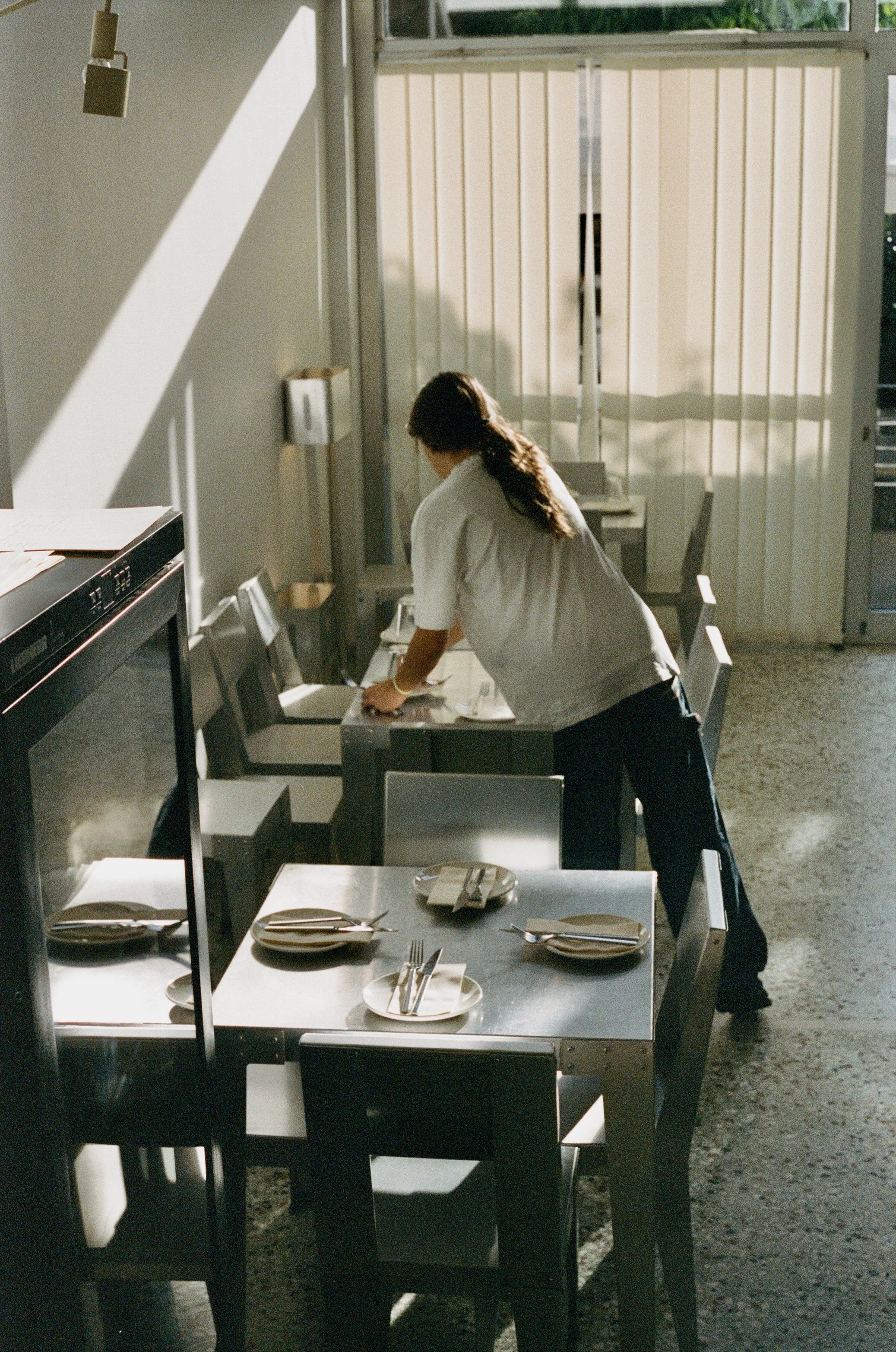 A woman arranging plates and utensils on a dining table in a sunlit room with chairs, a refrigerator, and vertical window blinds.