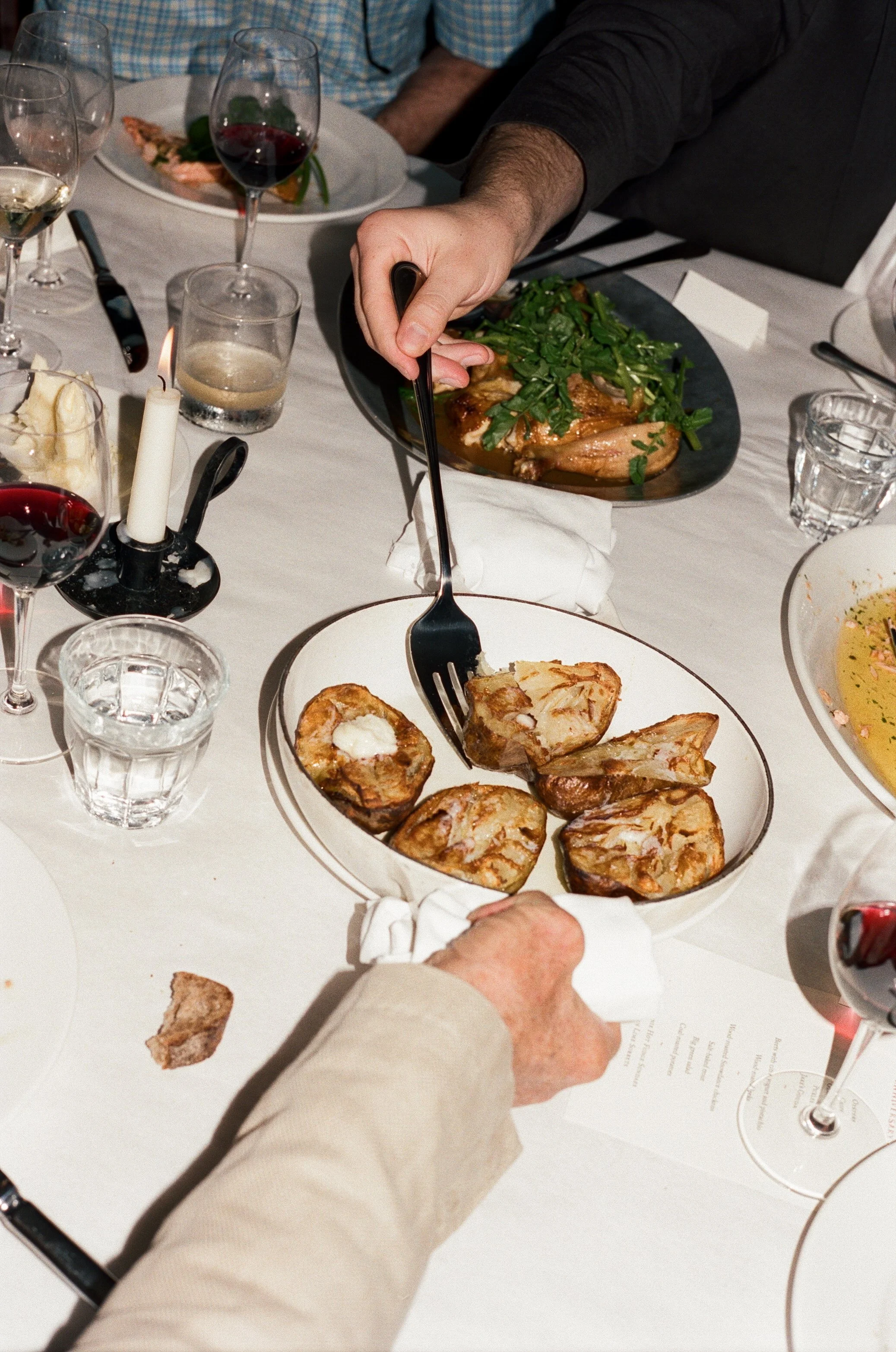 Person serving food at a dining table with plates of potatoes, meats, salads, wine glasses, and a lit candle.
