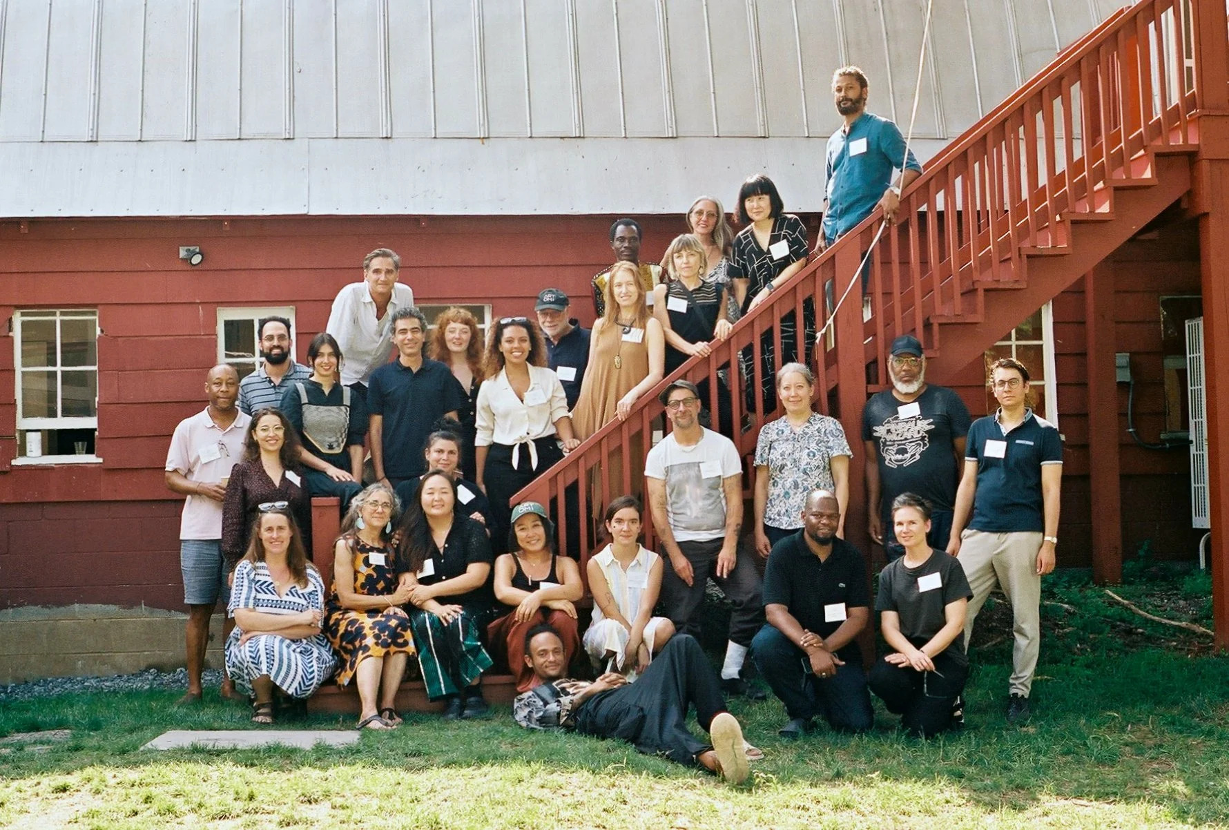 A diverse group of people posing outside in front of a building with a staircase, several people standing, sitting, and lying on the grass, smiling for the camera.