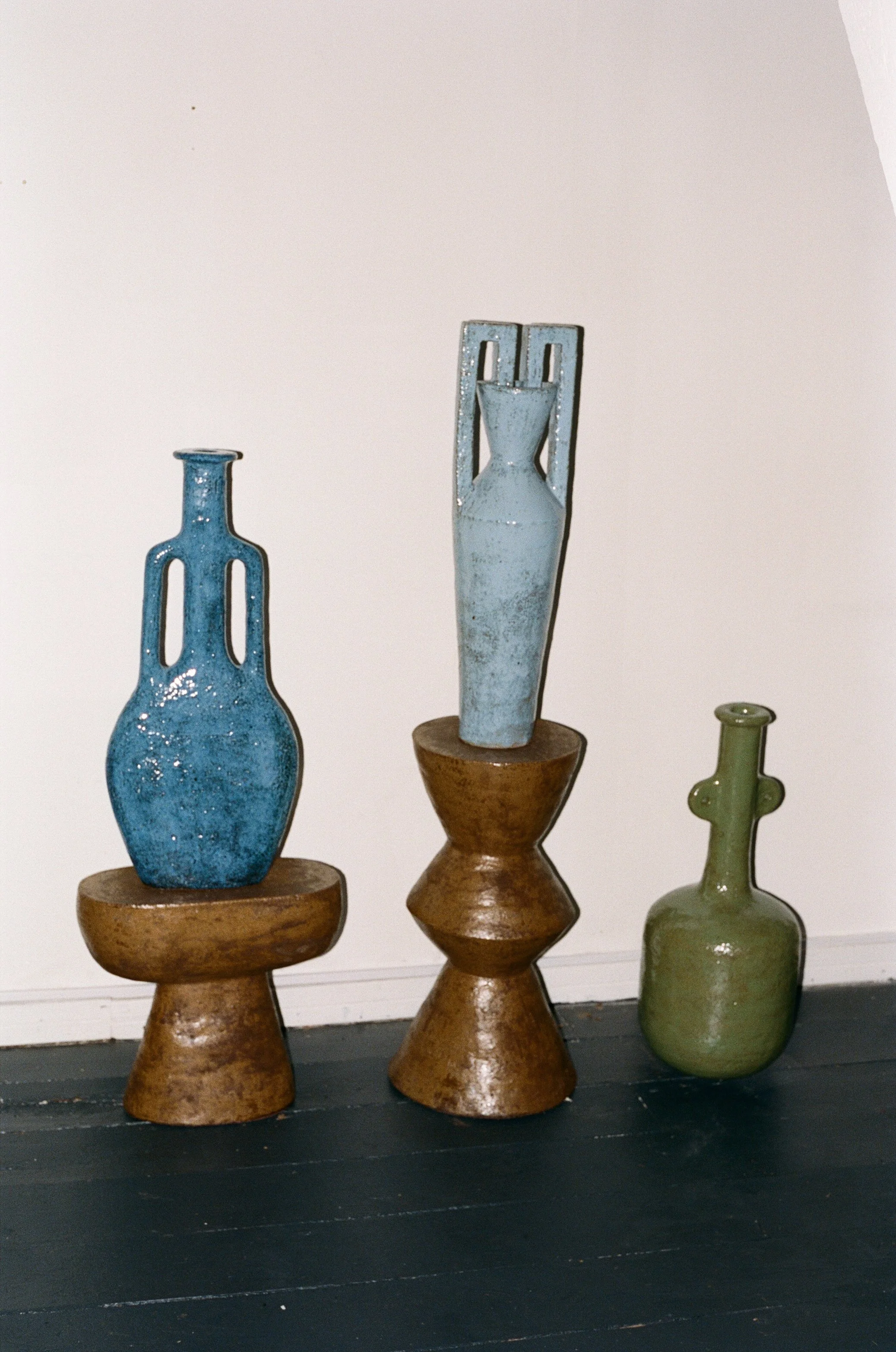 Three decorative ceramic vases with geometric shapes, each on wooden stands, arranged against a white wall.