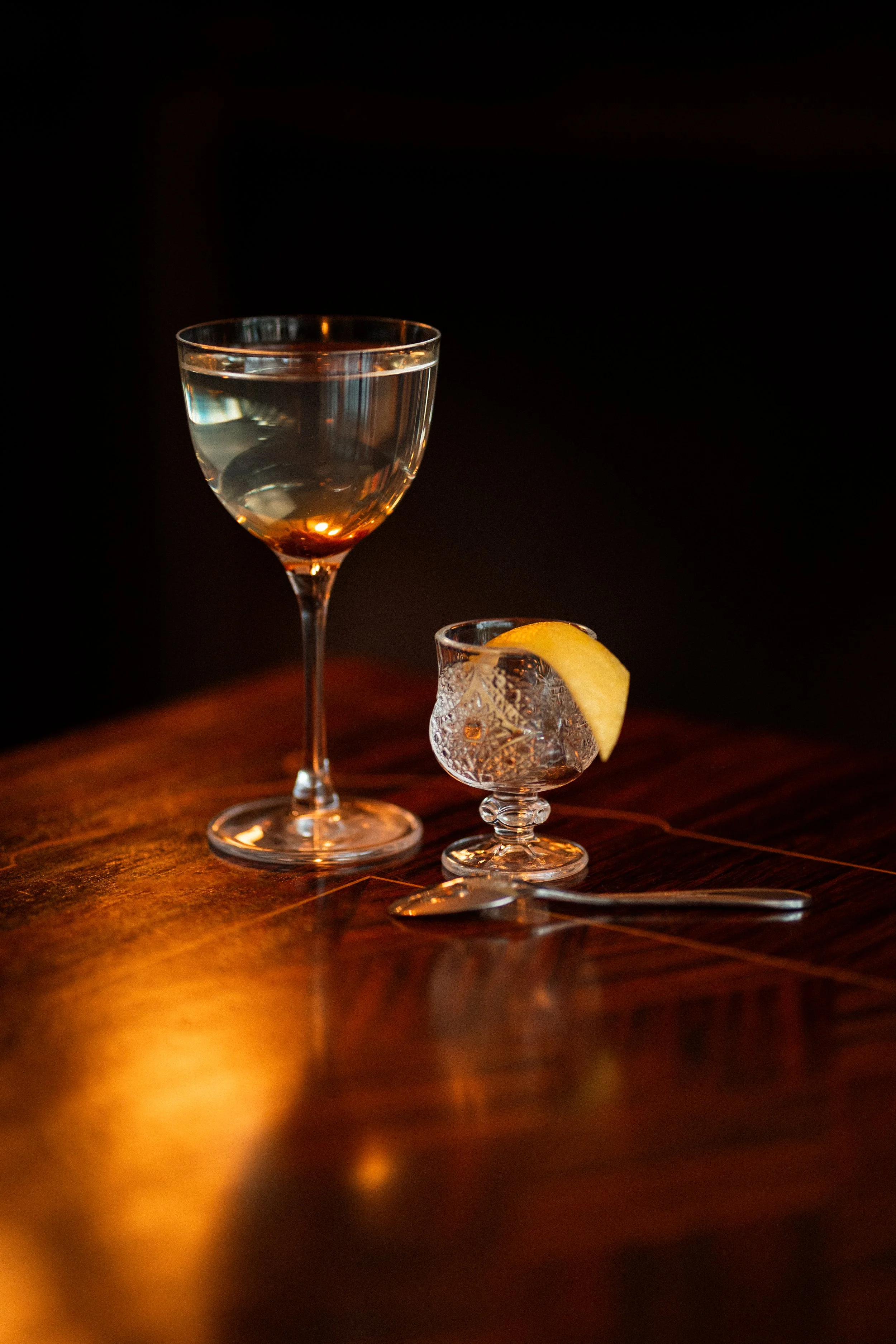A glass of white wine, a crystal glass with a lemon wedge, and a small silver spoon on a polished wooden surface with warm lighting.