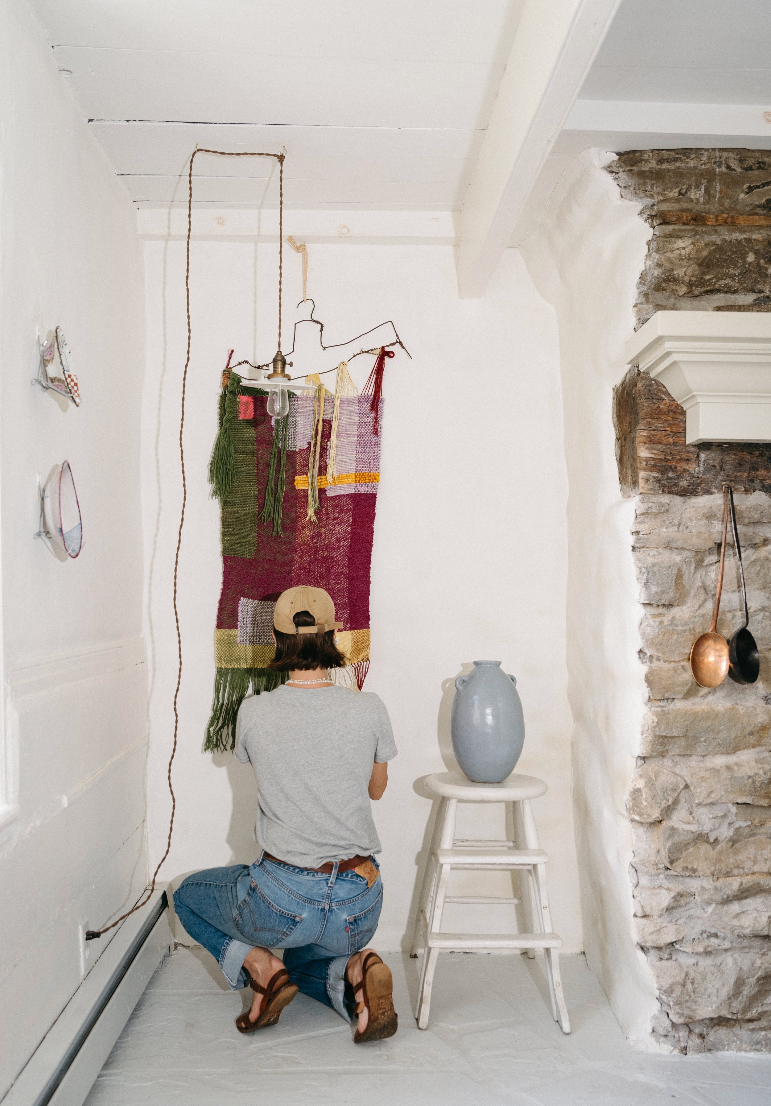 A person kneeling on the white floor, facing a white wall with a colorful woven textile hanging from a wire and a light bulb, with a large gray ceramic vase on a white stool beside them. The room has exposed stone on one wall and hanging copper cookw