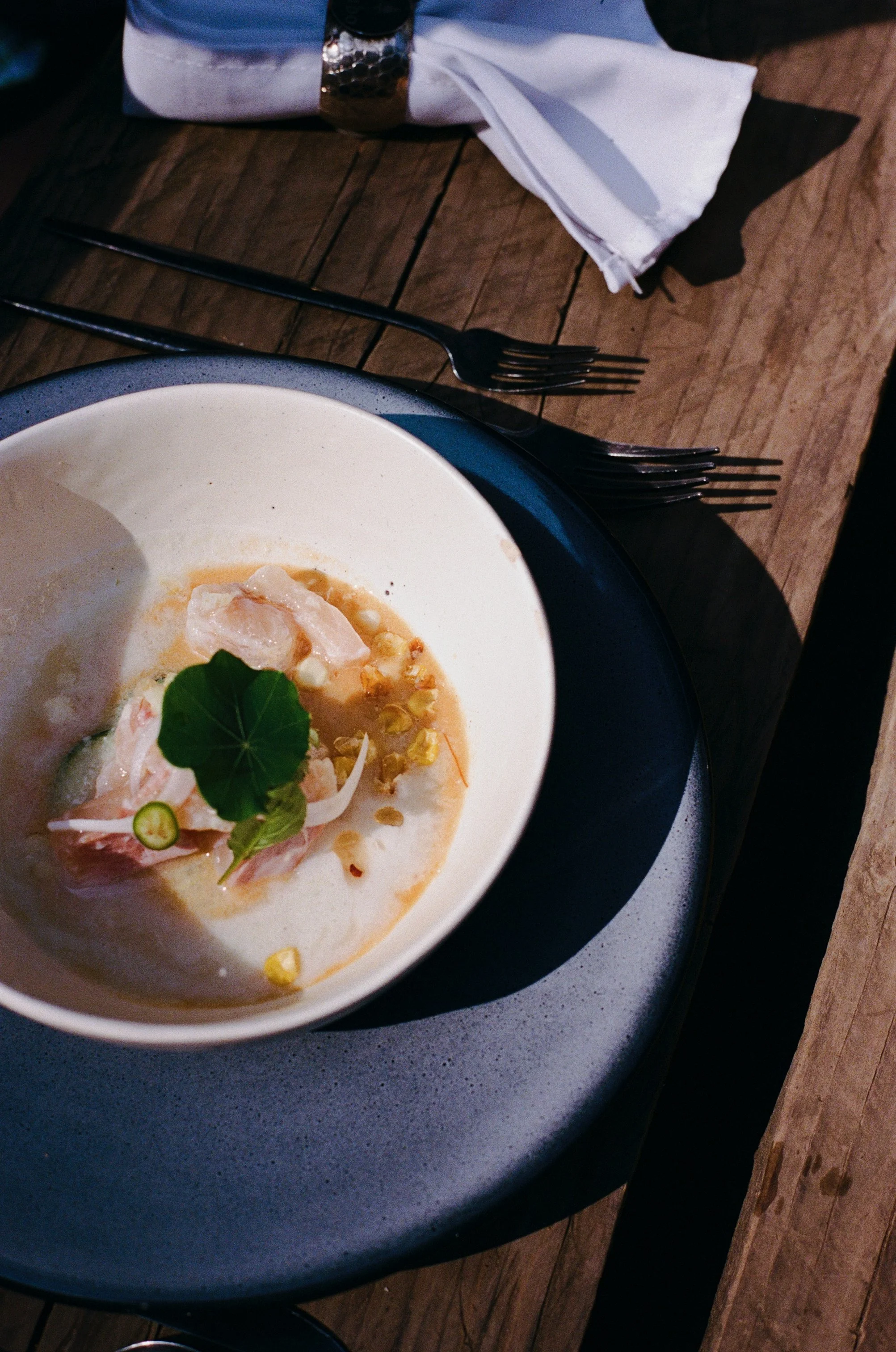 A dish of raw fish garnished with herbs and vegetables, served in a white bowl on a dark plate, on a rustic wooden table.