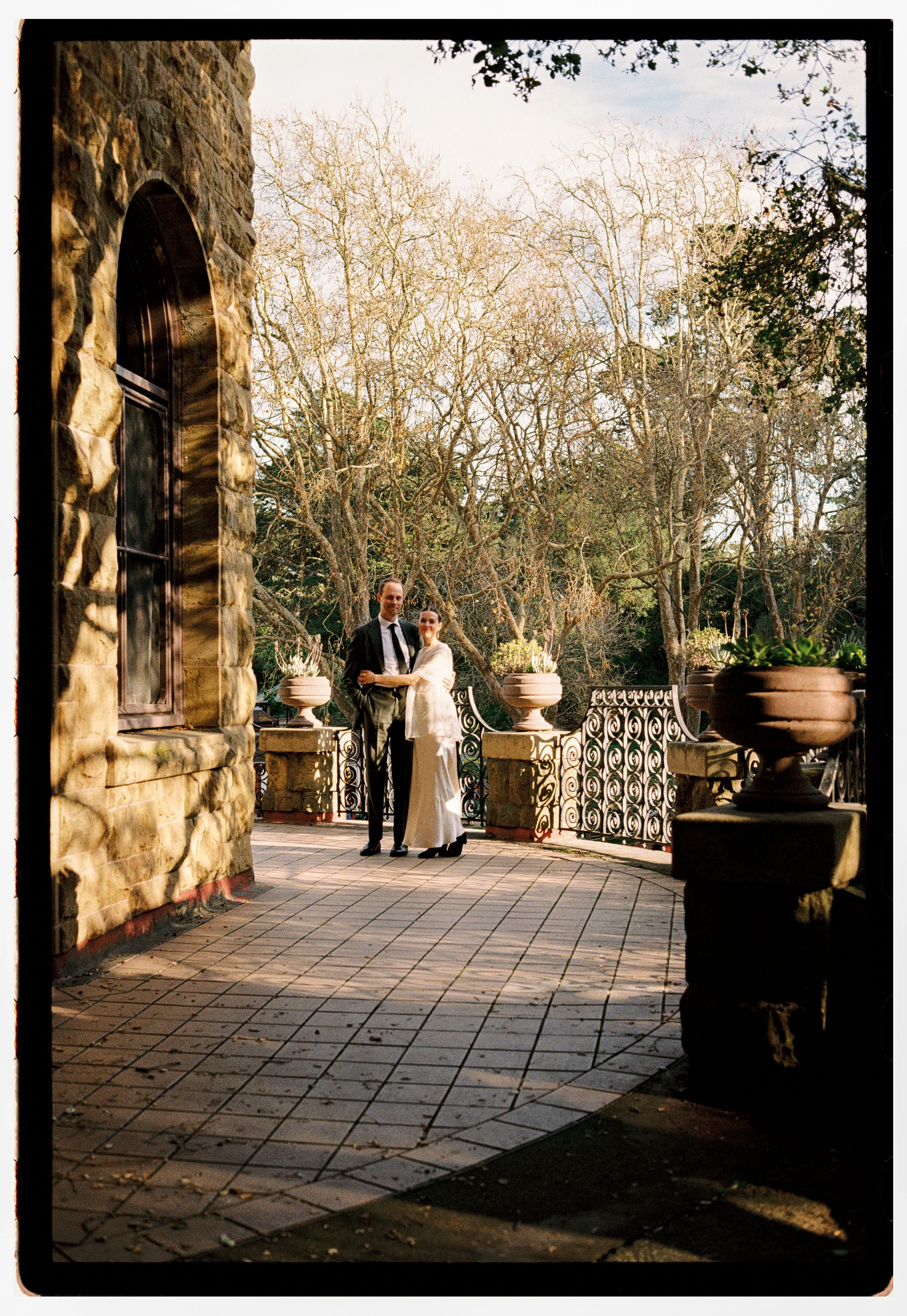 A couple dressed in wedding attire standing on a terrace with decorative railings and large potted plants, surrounded by leafless trees in a serene outdoor setting.