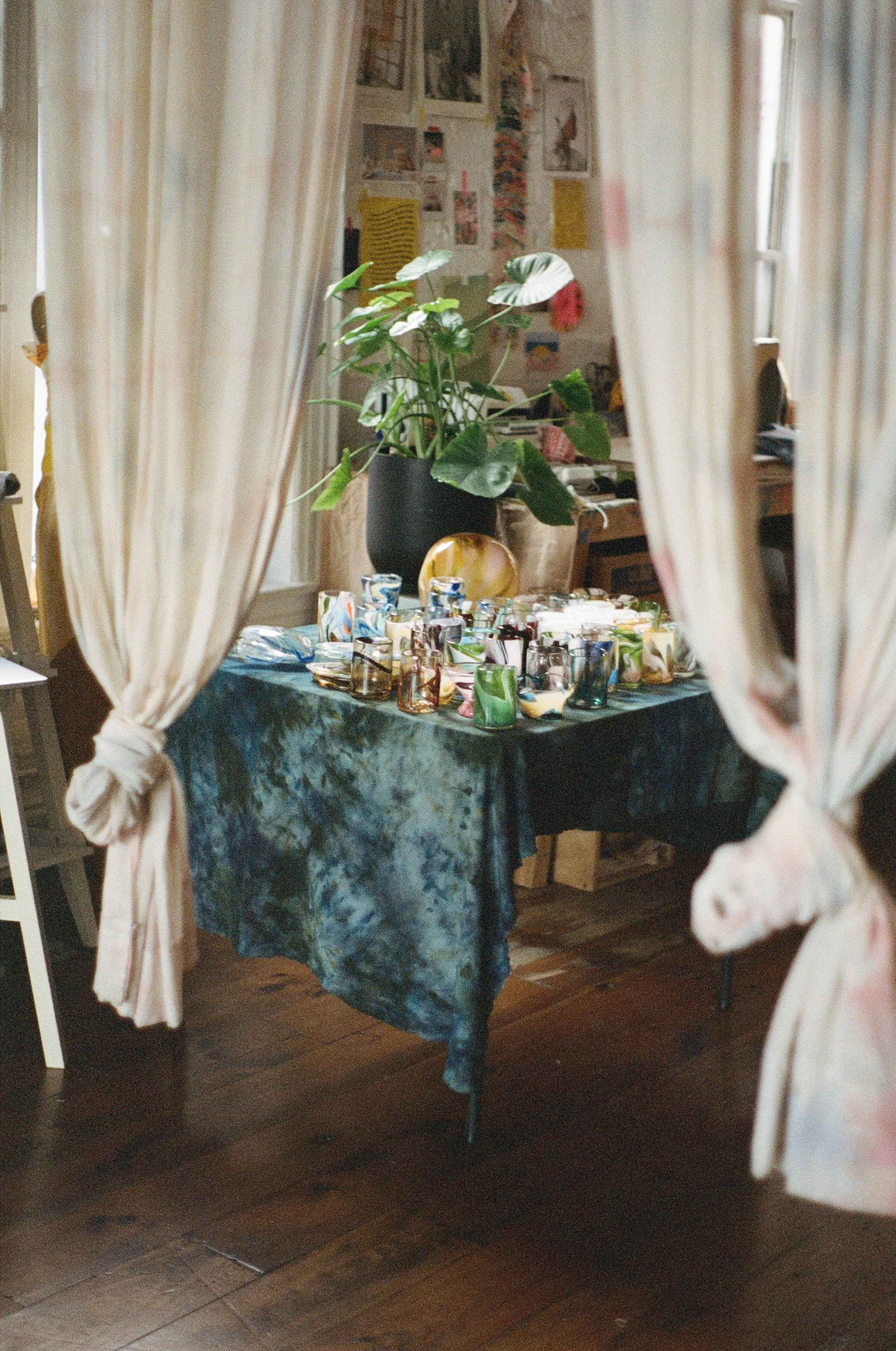 A table covered with a dark, tie-dye tablecloth, set with colorful glassware and ceramics, seen through a gap in floral curtains, with a large potted plant in the background.