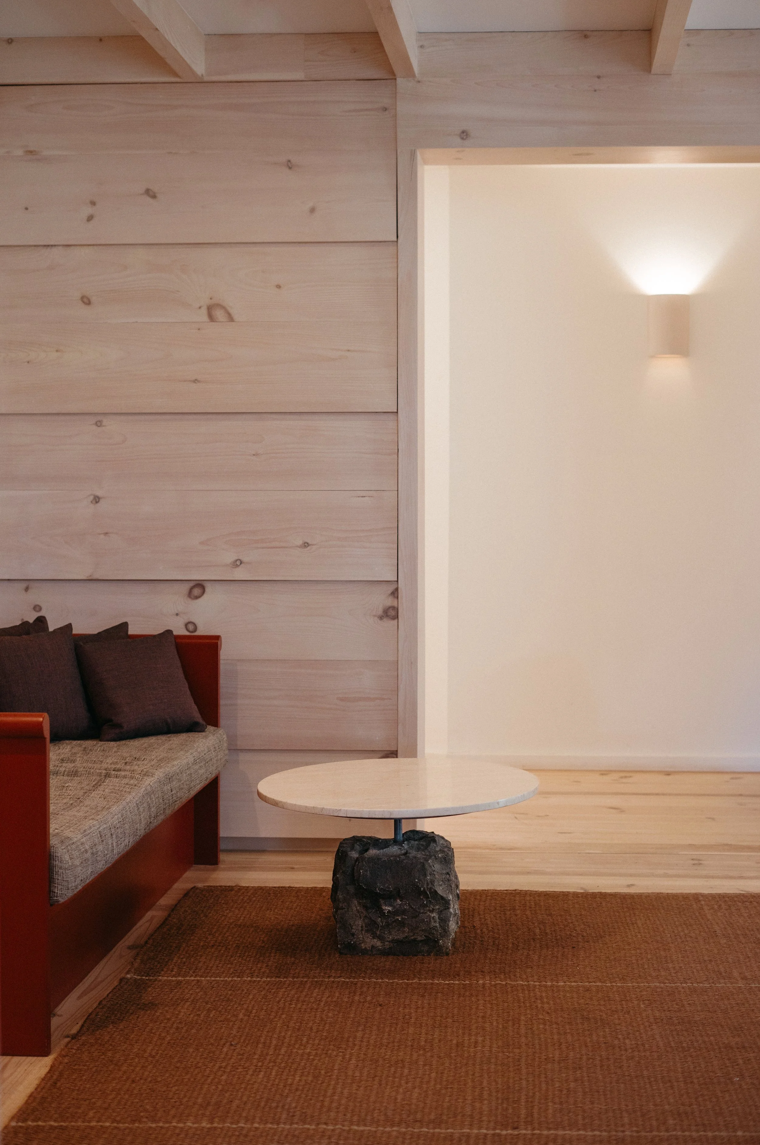 Living room with wooden paneled wall, small round table with stone base, couch with dark pillows, and a modern wall-mounted light fixture.