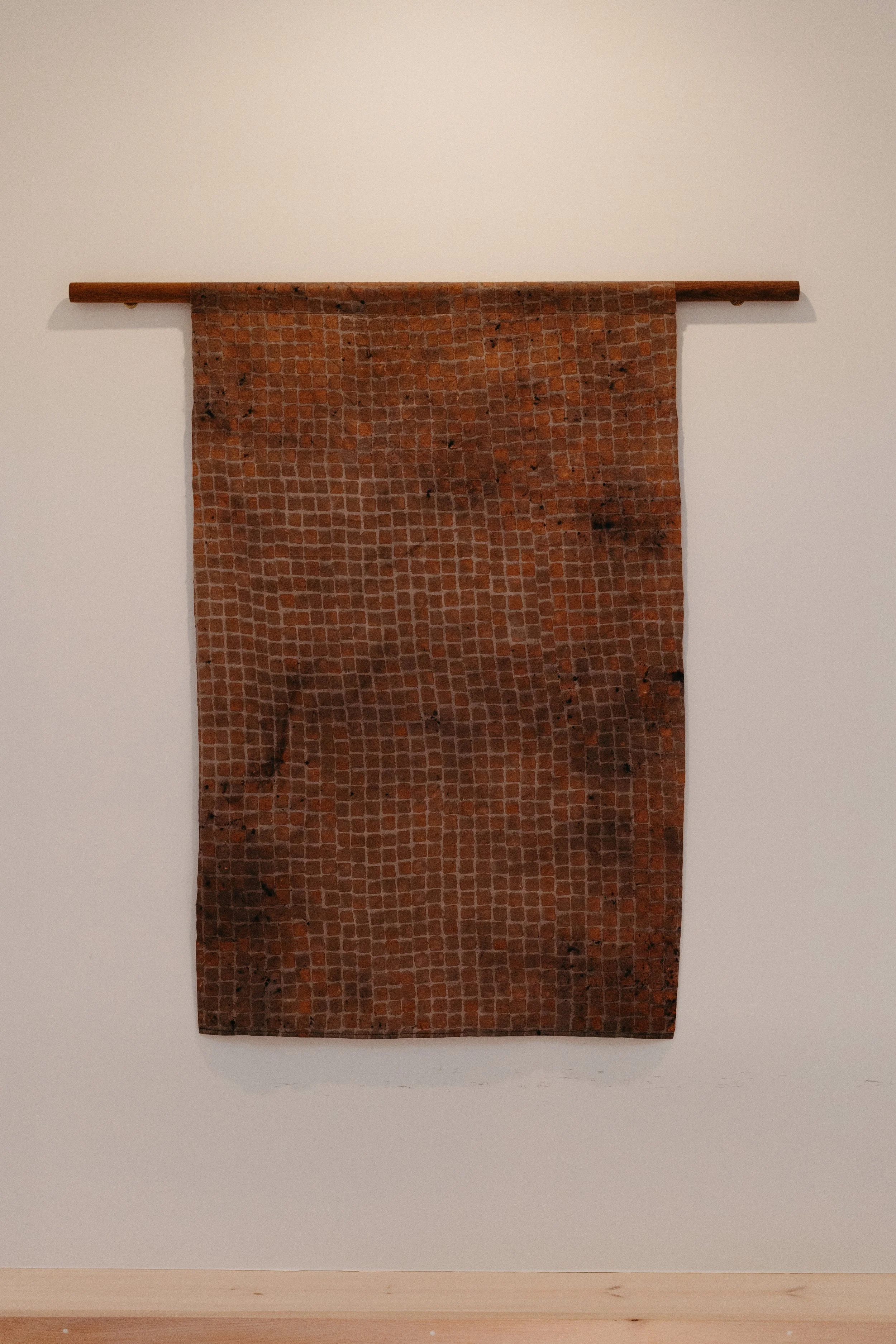 A textile artwork hanging on a white gallery wall, attached to a wooden rod at the top.