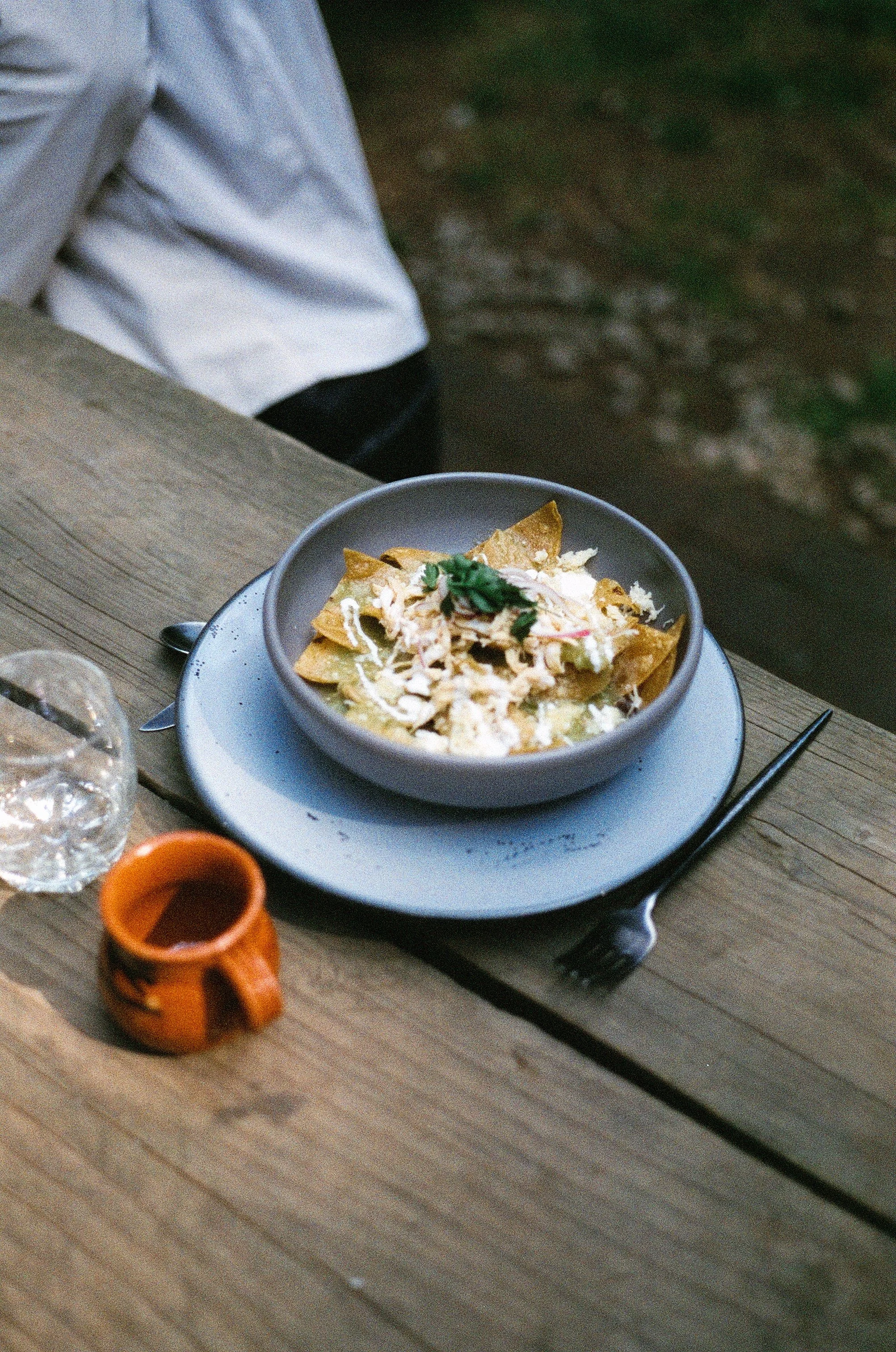 A bowl of chilaquiles with shredded chicken, crema, and a garnish of cilantro on a wooden table, with a small glass of water and a tiny clay cup nearby.