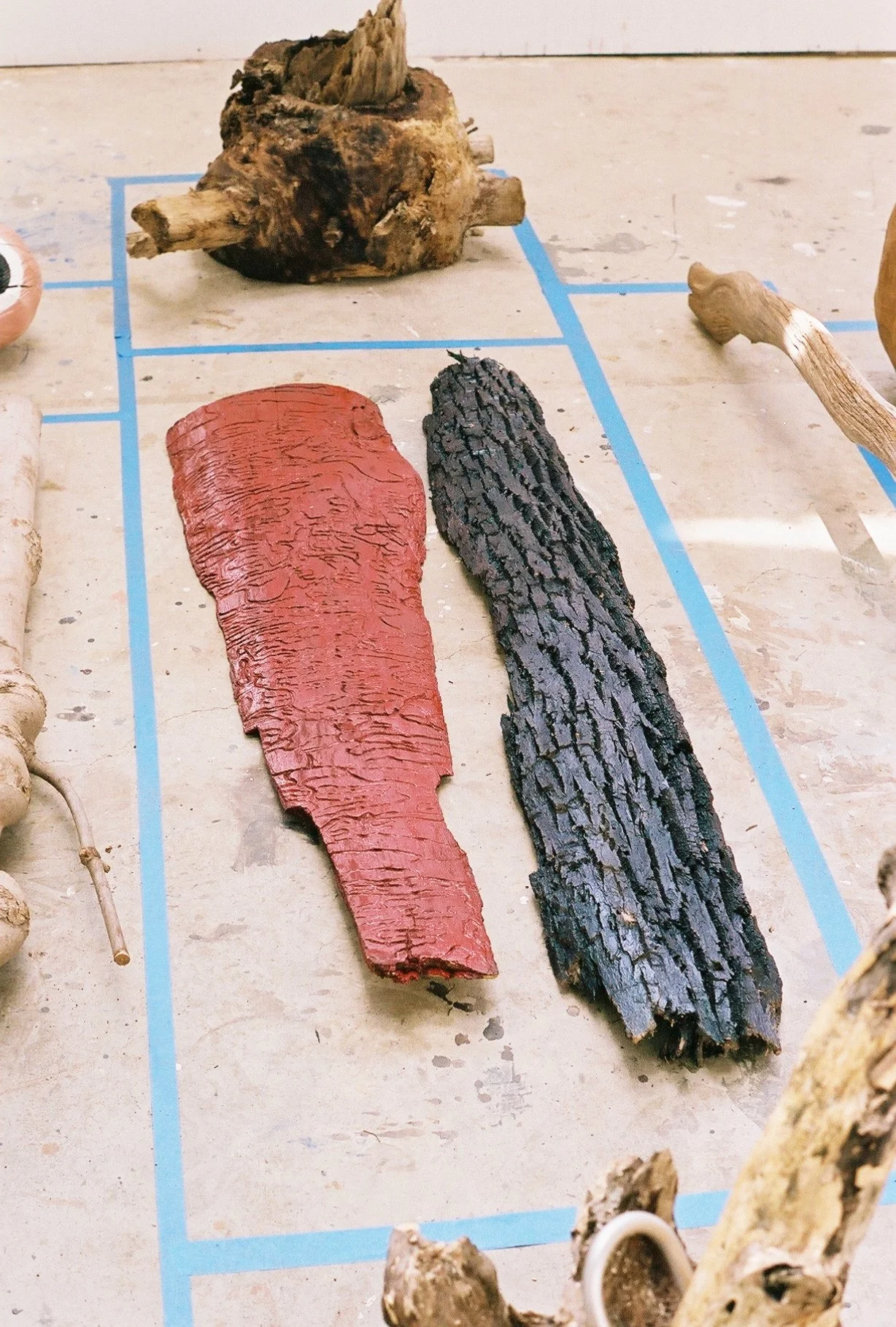 Two pieces of wood, one red and one blackened with bark, are placed on a blue grid on a concrete surface. The red piece appears painted or treated, while the black piece looks burnt or charred. Other pieces of wood are partially visible around the gr