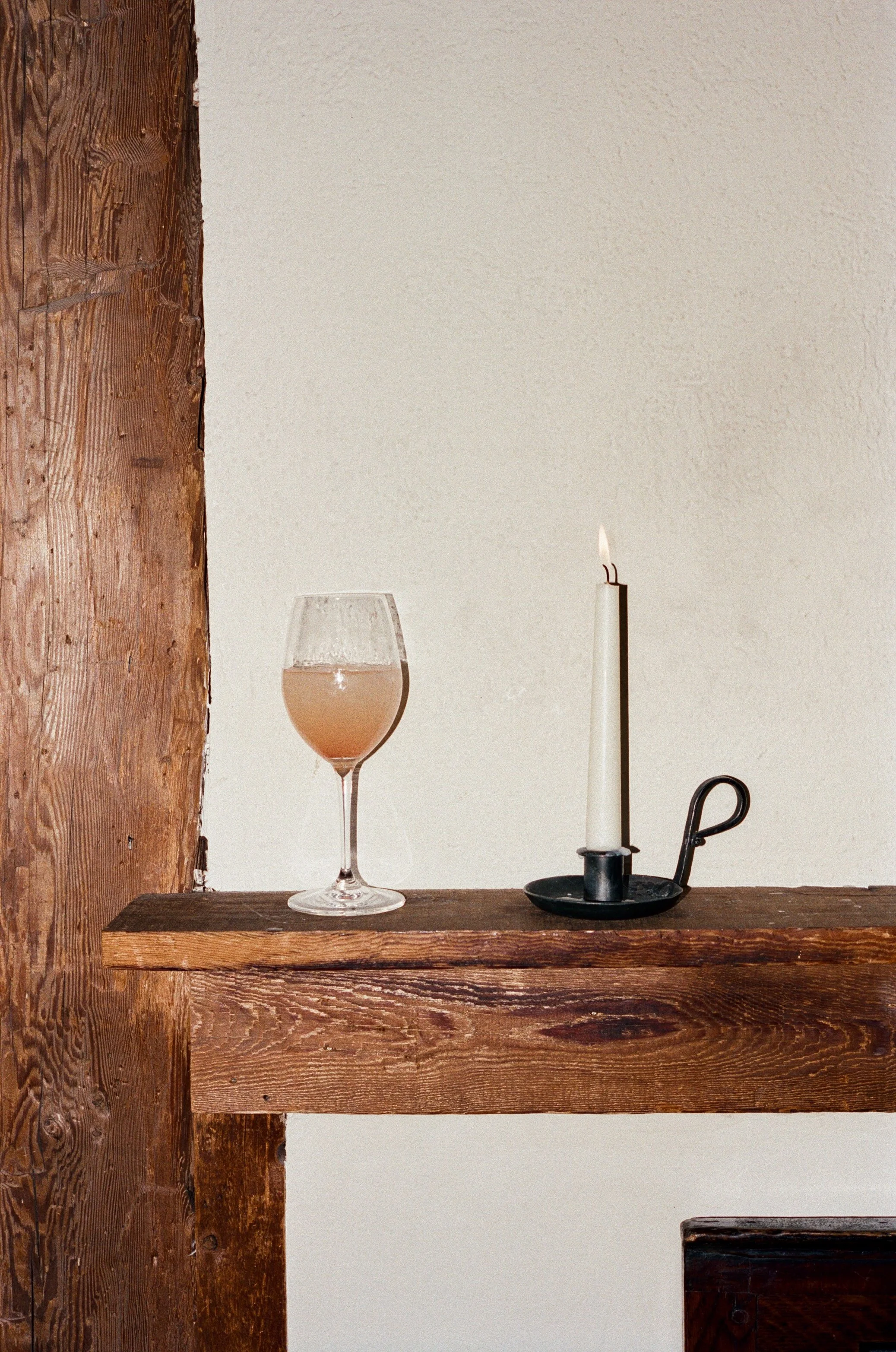 A wooden shelf with a glass of pink cocktail and a lit white candle in a black holder against a plain wall.