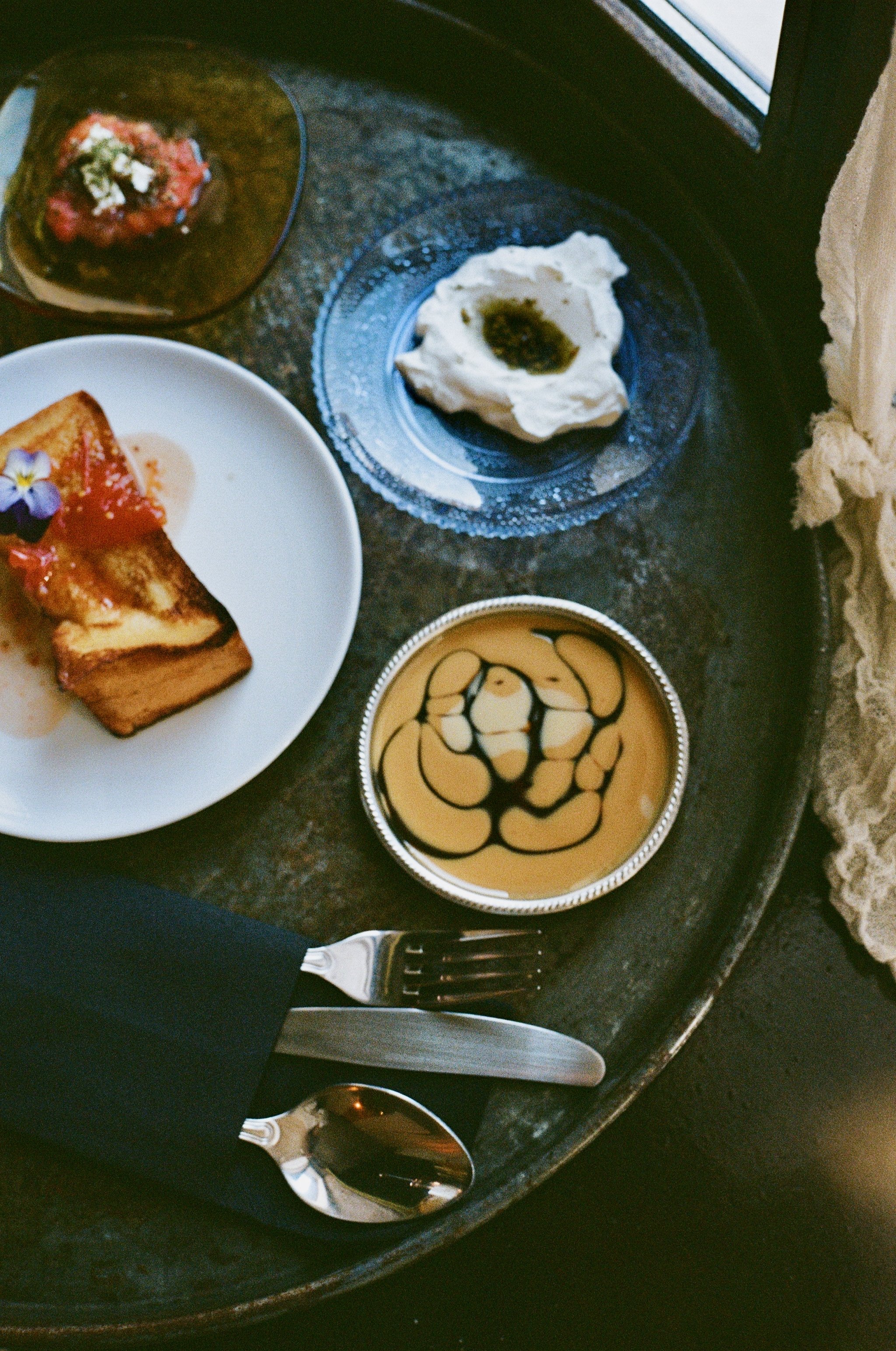 A tray with various desserts, including French toast with edible flower, whipped cream with syrup, a caramel drink with syrup design, and a small bowl of green and red sauce.
