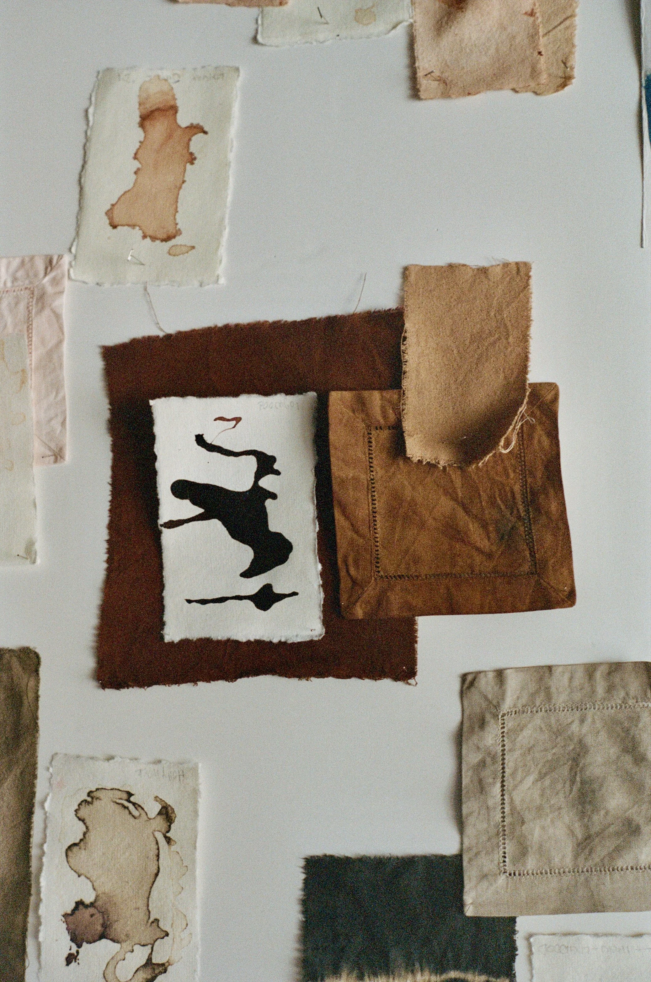 A collage of fabric and paper swatches on a white surface, including various textures and colors such as brown, beige, black, and cream, with some pieces featuring ink or paint designs.