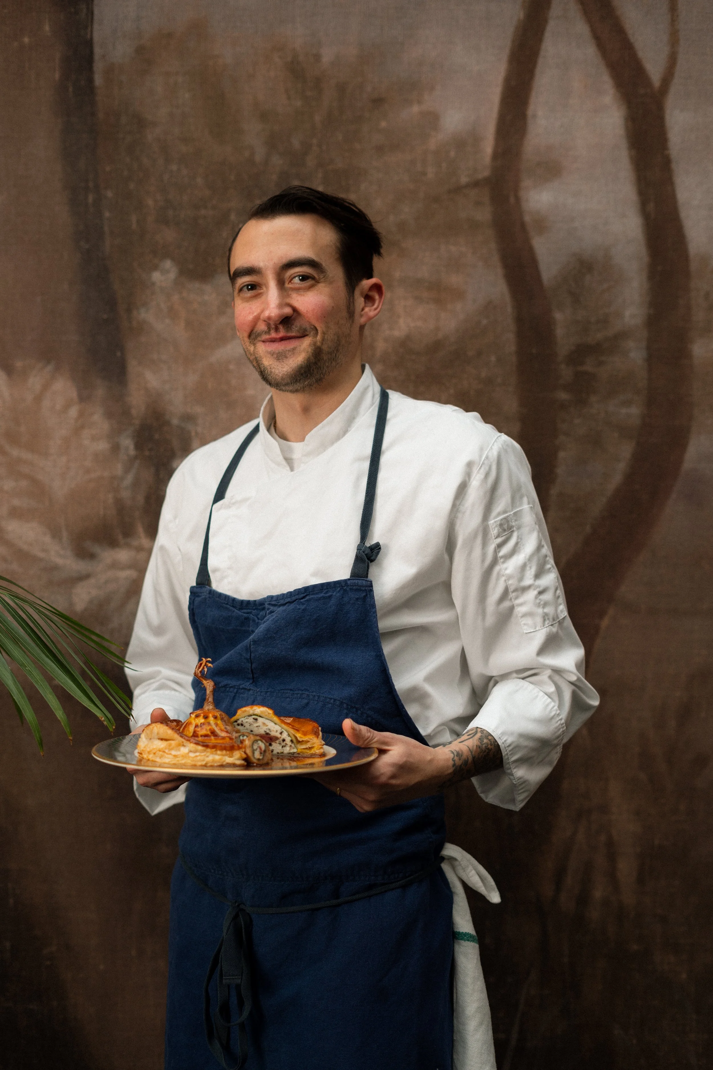 A chef in a white uniform and navy apron holding a plate of prepared dishes, standing in front of a brown textured background.