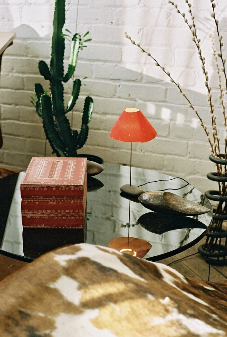 Decorative table with a small red lamp, a cactus plant in a black pot, a vintage red woodworking ruler box, smooth stones, and dried branches against a white brick wall.