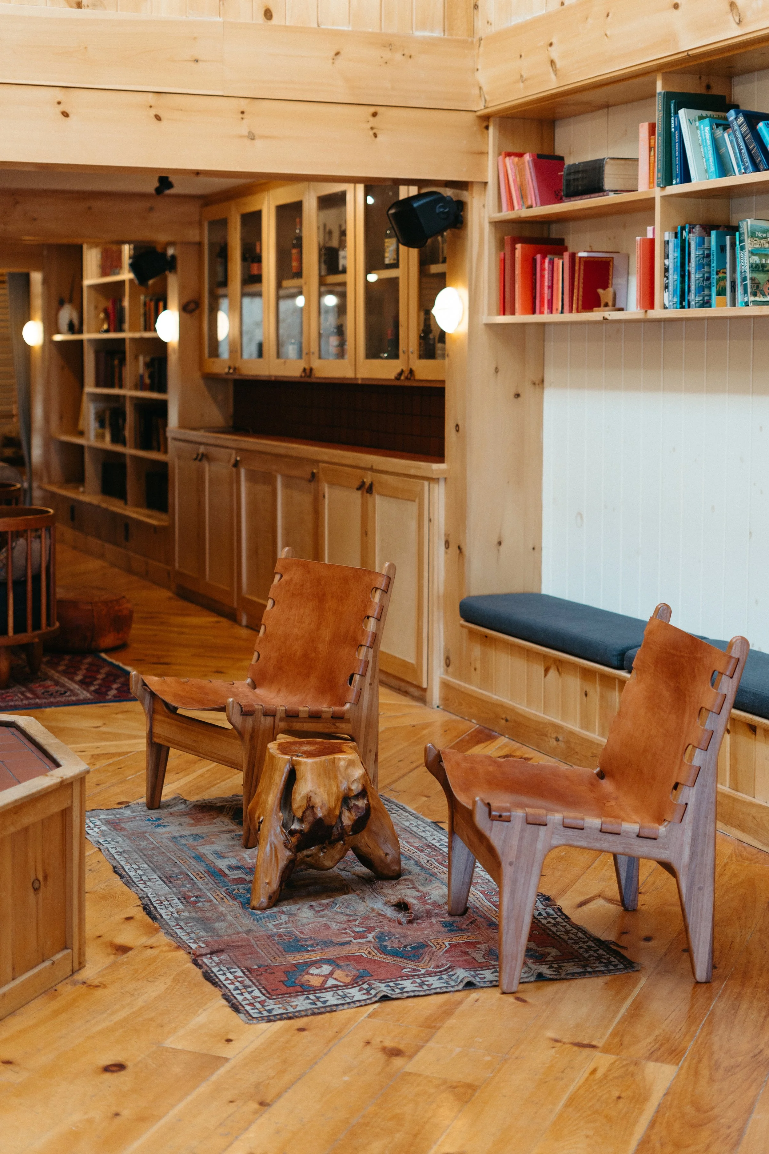 A cozy interior with wooden walls and flooring, featuring two leather chairs, a small wooden table, a built-in cushioned bench, and bookshelves filled with colorful books.