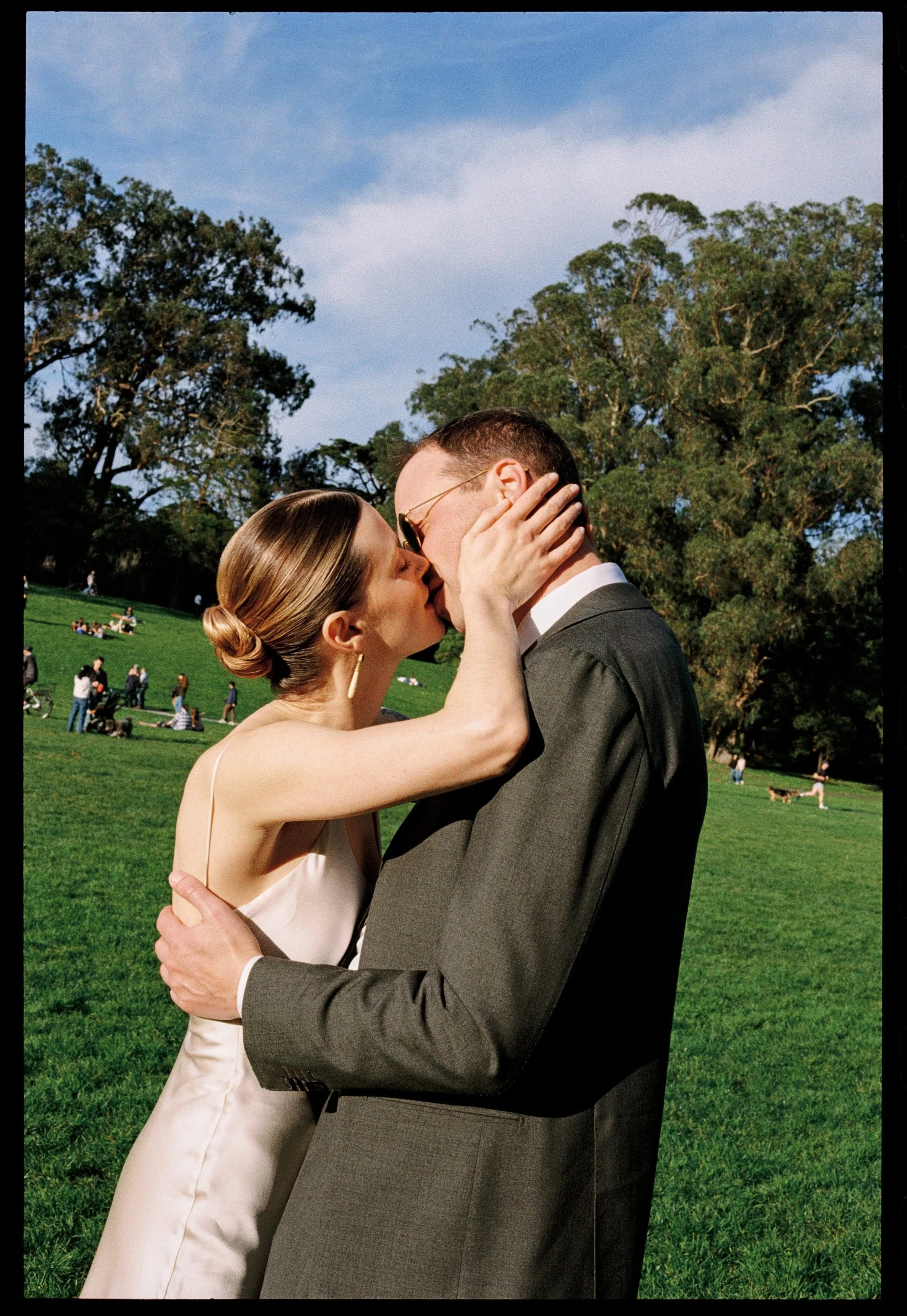 A couple sharing a kiss in Golden Gate Park with green grass and trees, people lounging, and dogs in the background on a sunny day.
