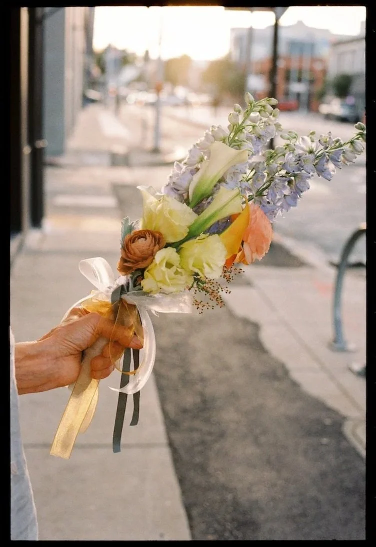 Person holding a small bouquet of mixed flowers on a city sidewalk during sunset.