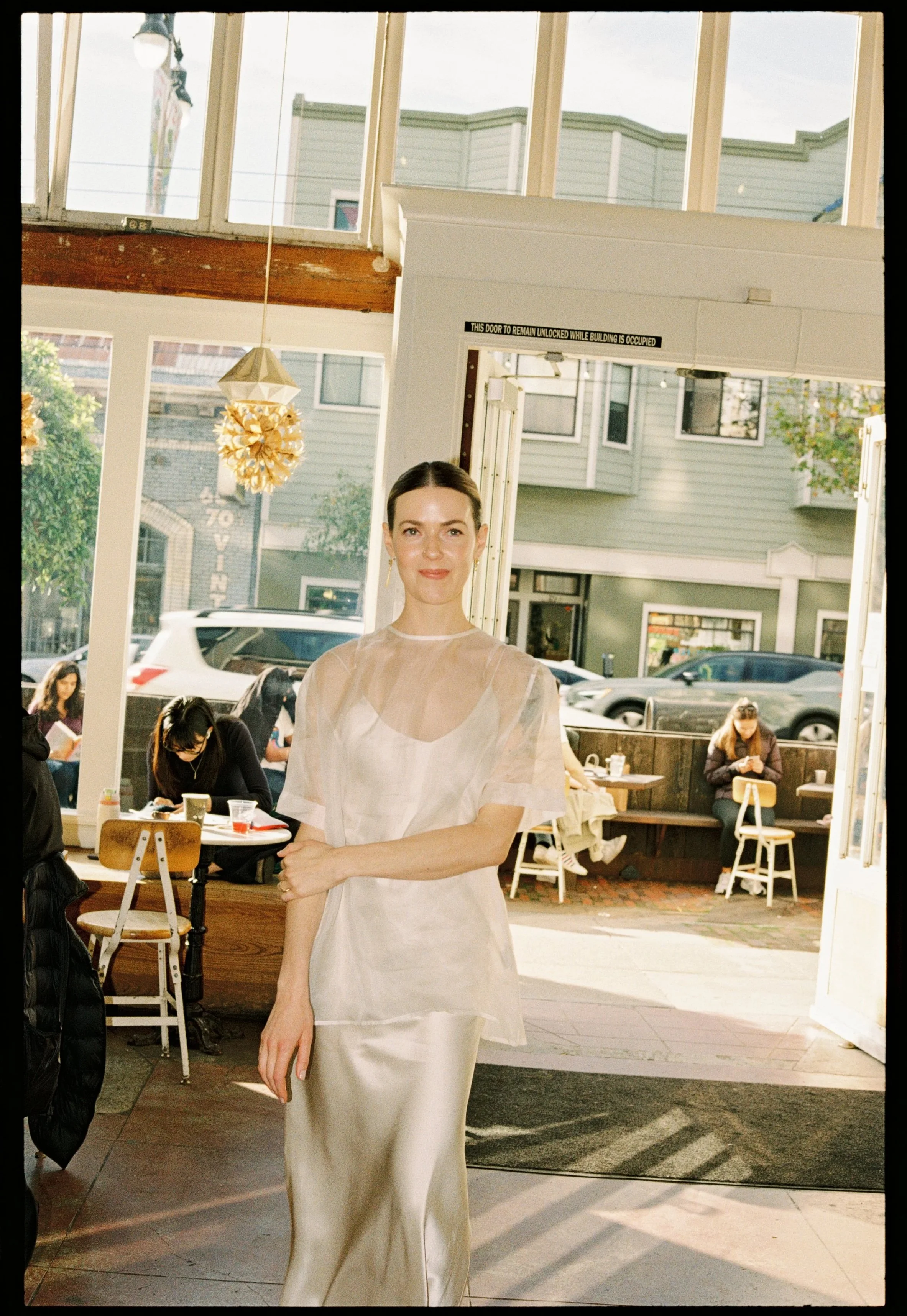 A woman standing inside a cafe or restaurant near the entrance, wearing a cream-colored silk dress with a sheer overlay, smiling at the camera. Through the open door and large windows, parked cars and people sitting outside are visible.