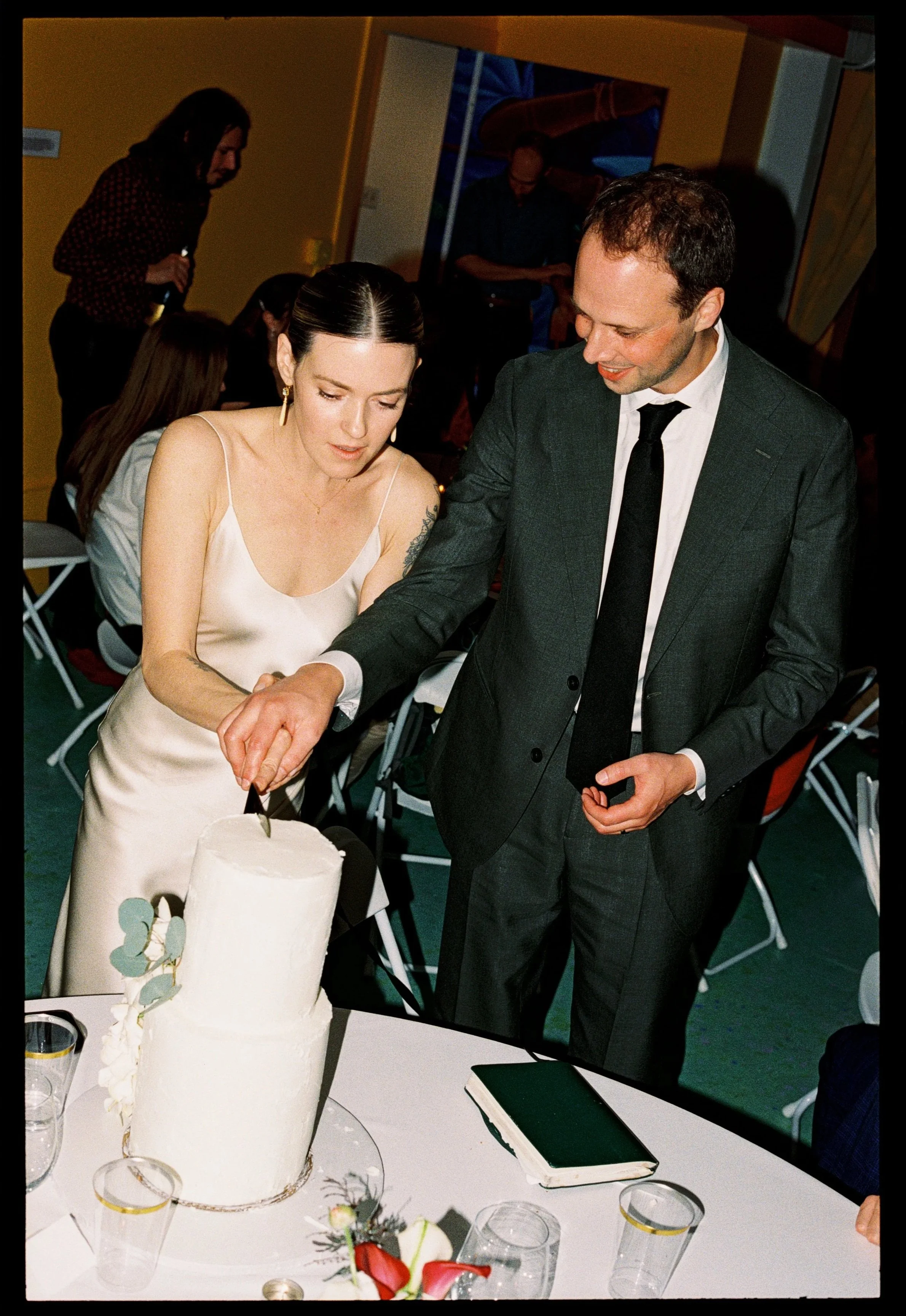 A woman in a white dress and a man in a suit are cutting a white wedding cake together at a celebration. There are glasses and flowers on the table in front of them, and other people are visible in the background.