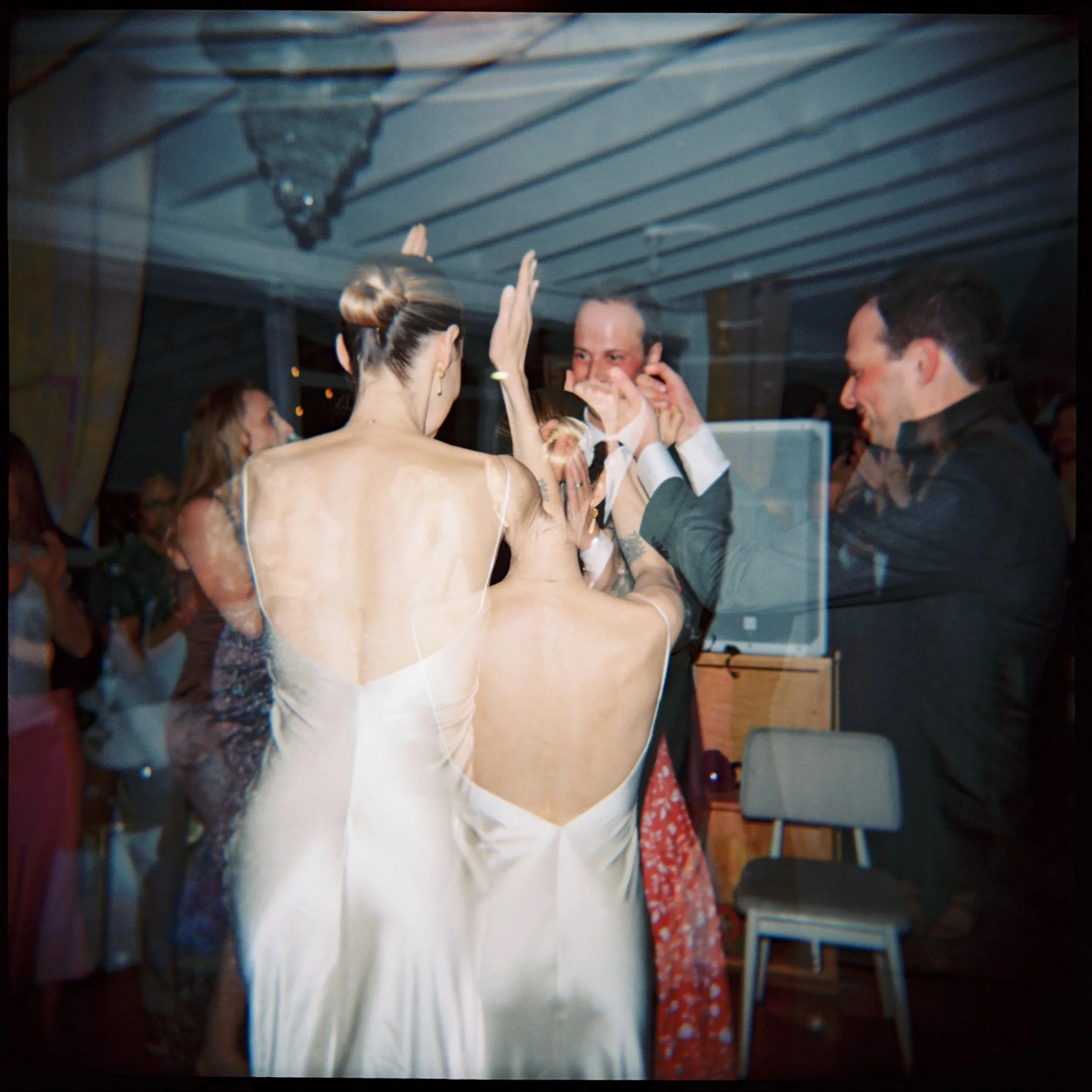People dancing and celebrating at a wedding reception, with women in elegant dresses and men in suits, inside a decorated venue.