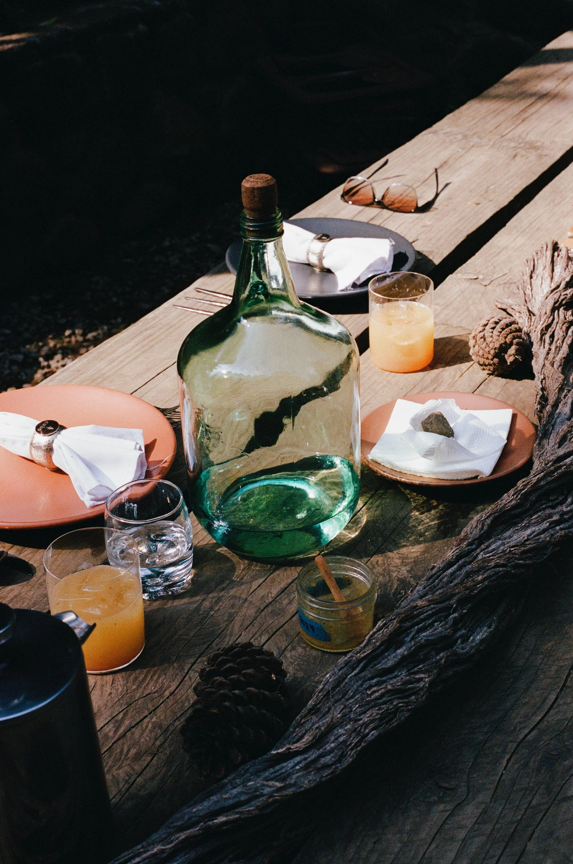 Outdoor wooden table with a large green glass jug, orange plates with napkins and cutlery, glasses of water and juice, small dish with a rock, sunglasses, and pinecones.