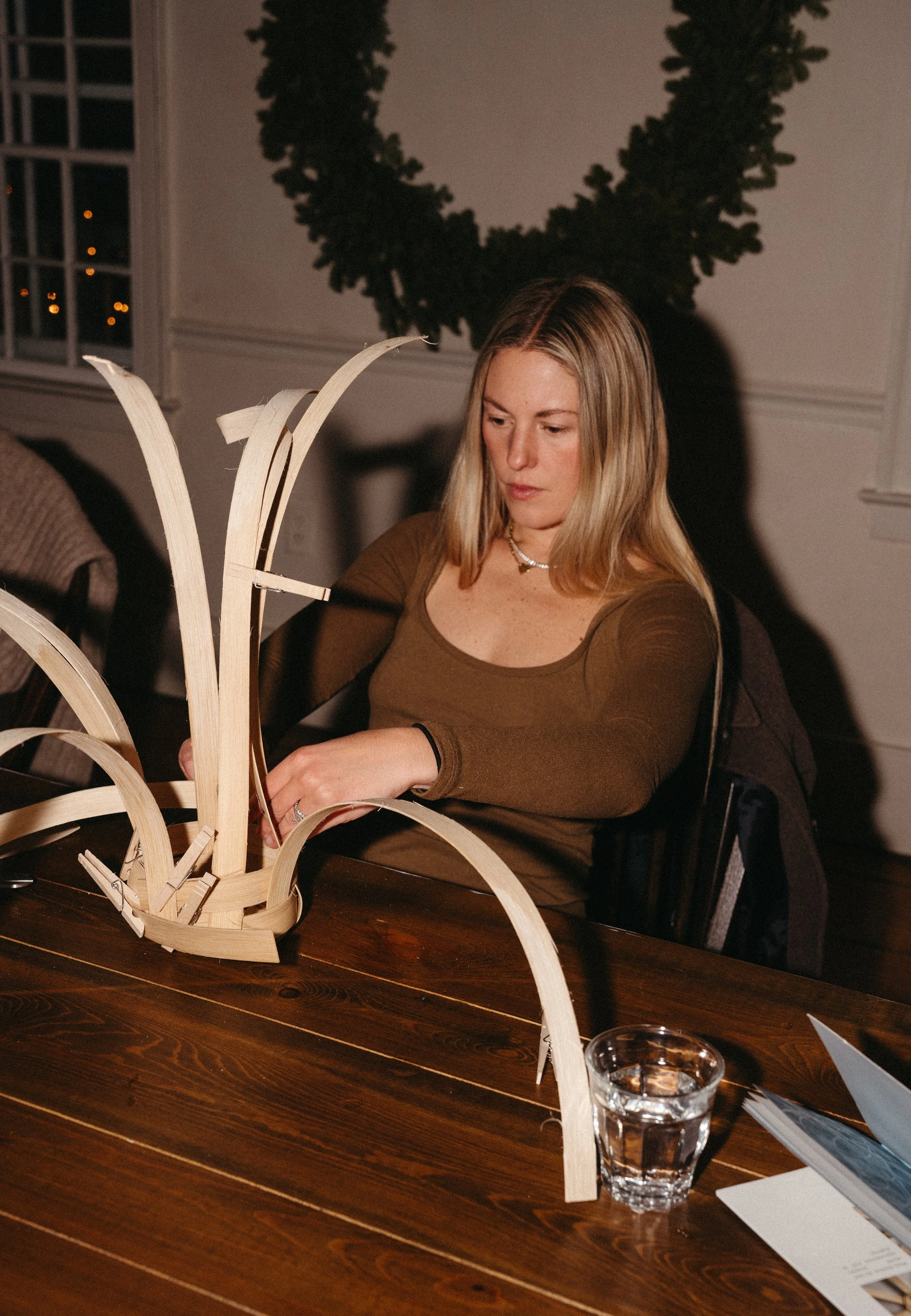 A woman with blonde hair decorating a wooden reindeer sculpture at a dinner table in a festive setting with a wreath hanging on the wall behind her.