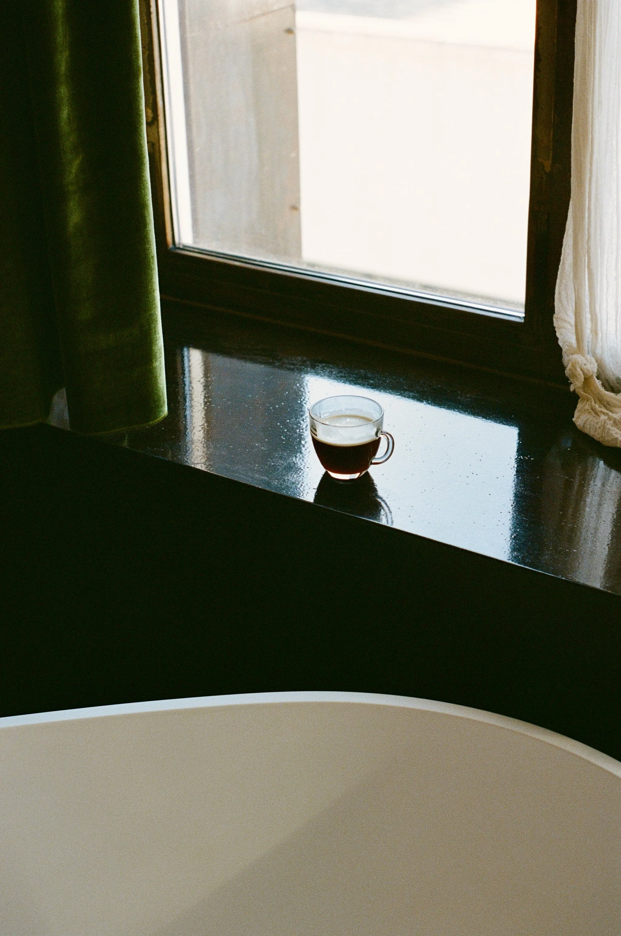 A small glass cup filled with dark coffee placed on a glossy black windowsill near a window with light curtains.