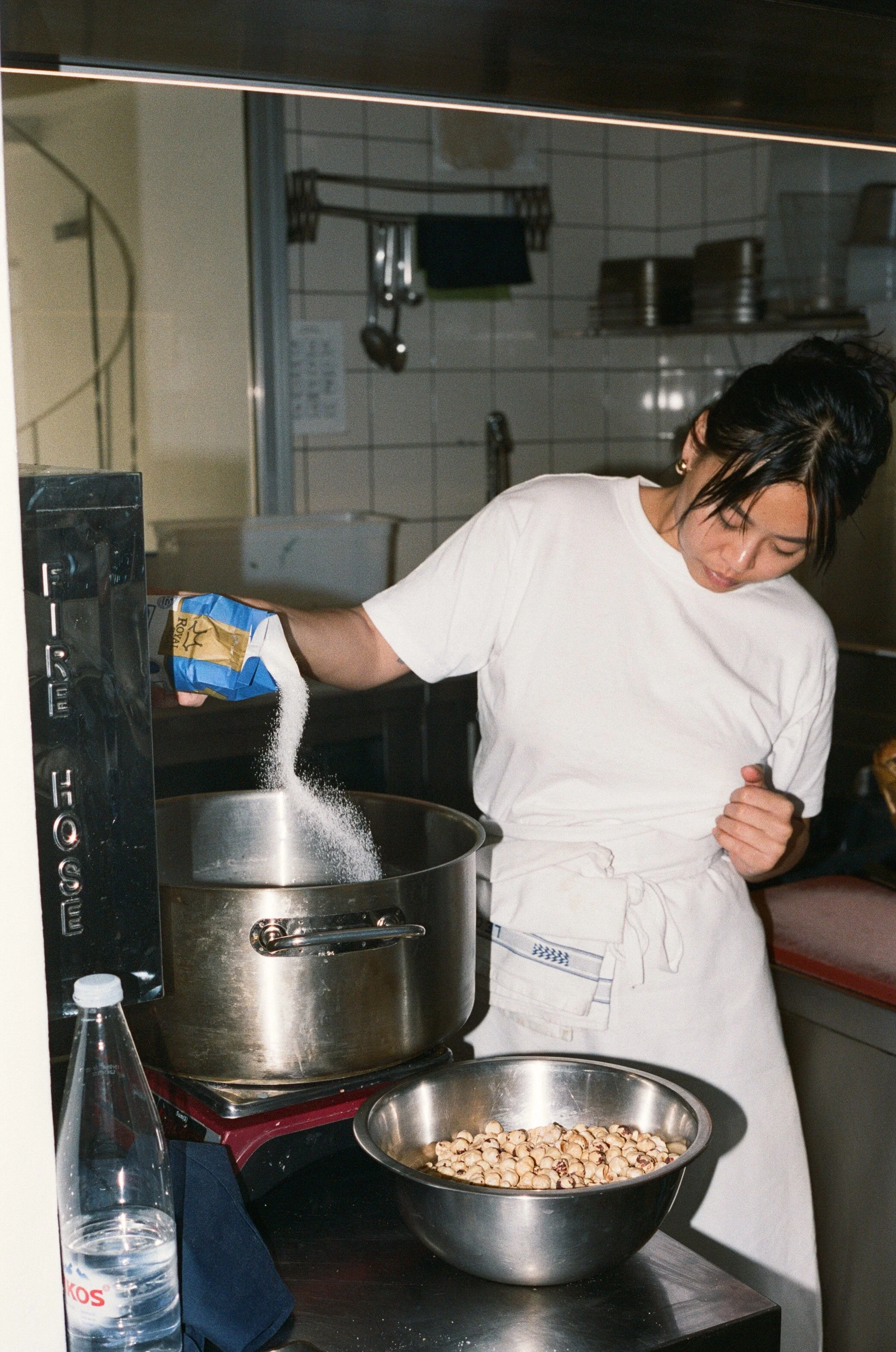 A woman is pouring sugar into a pot in a kitchen. A metal bowl with hazelnuts sits on the counter nearby.
