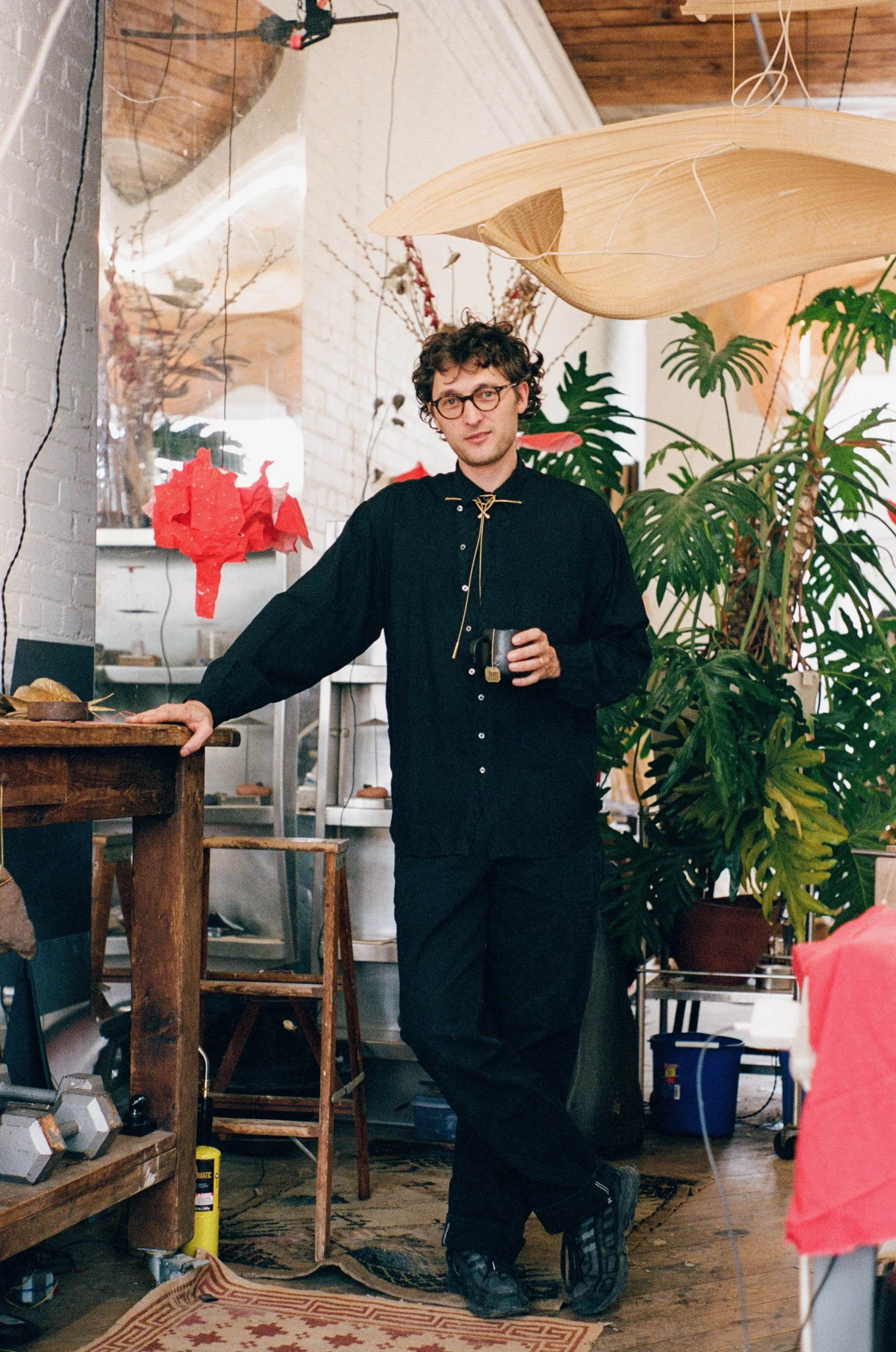 A man with curly hair, glasses, and a black shirt standing indoors, holding a small cup, with large green plants, wooden furniture, and decorative paper art in the background.