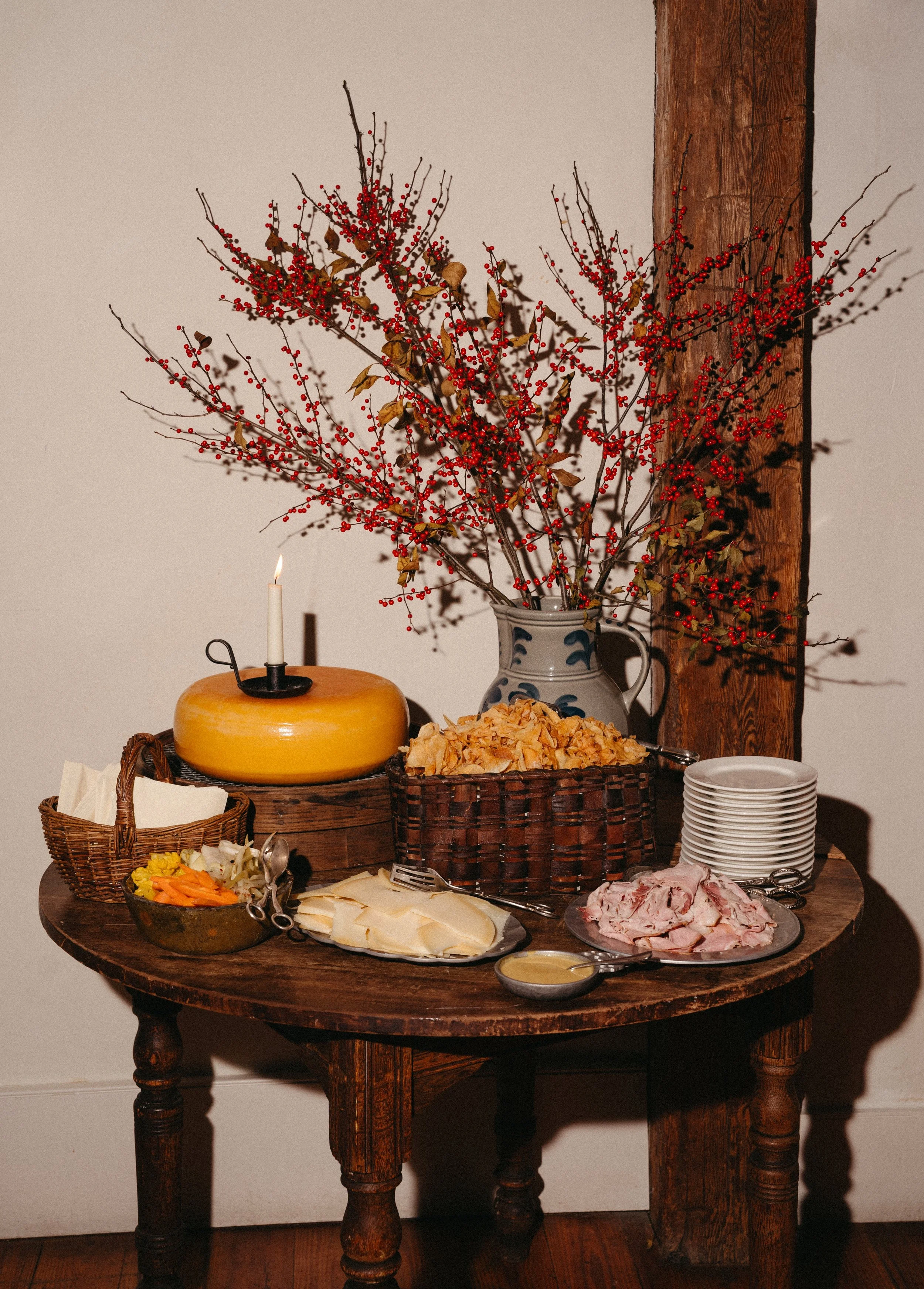 A wooden table with a large bouquet of red berries and branches in a ceramic pitcher, a round yellow cheese with a candle on top, a basket of potato chips, a plate of sliced cheese, a plate of sliced ham, a small bowl of yellow sauce, a side dish wit