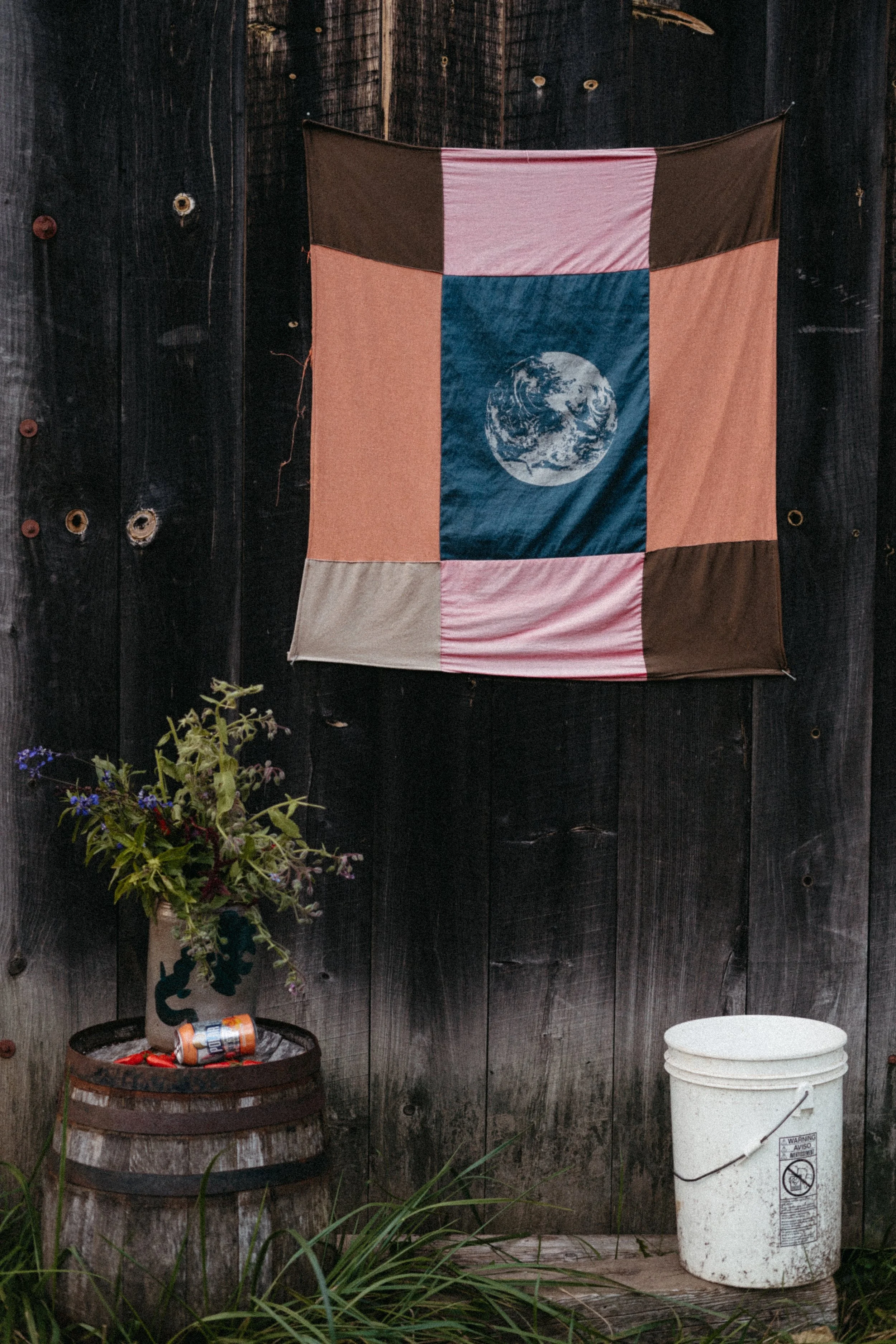 Colorful quilt with an image of Earth hanging on a dark wooden wall. A flower arrangement in a pottery vase and a white bucket are on the ground.