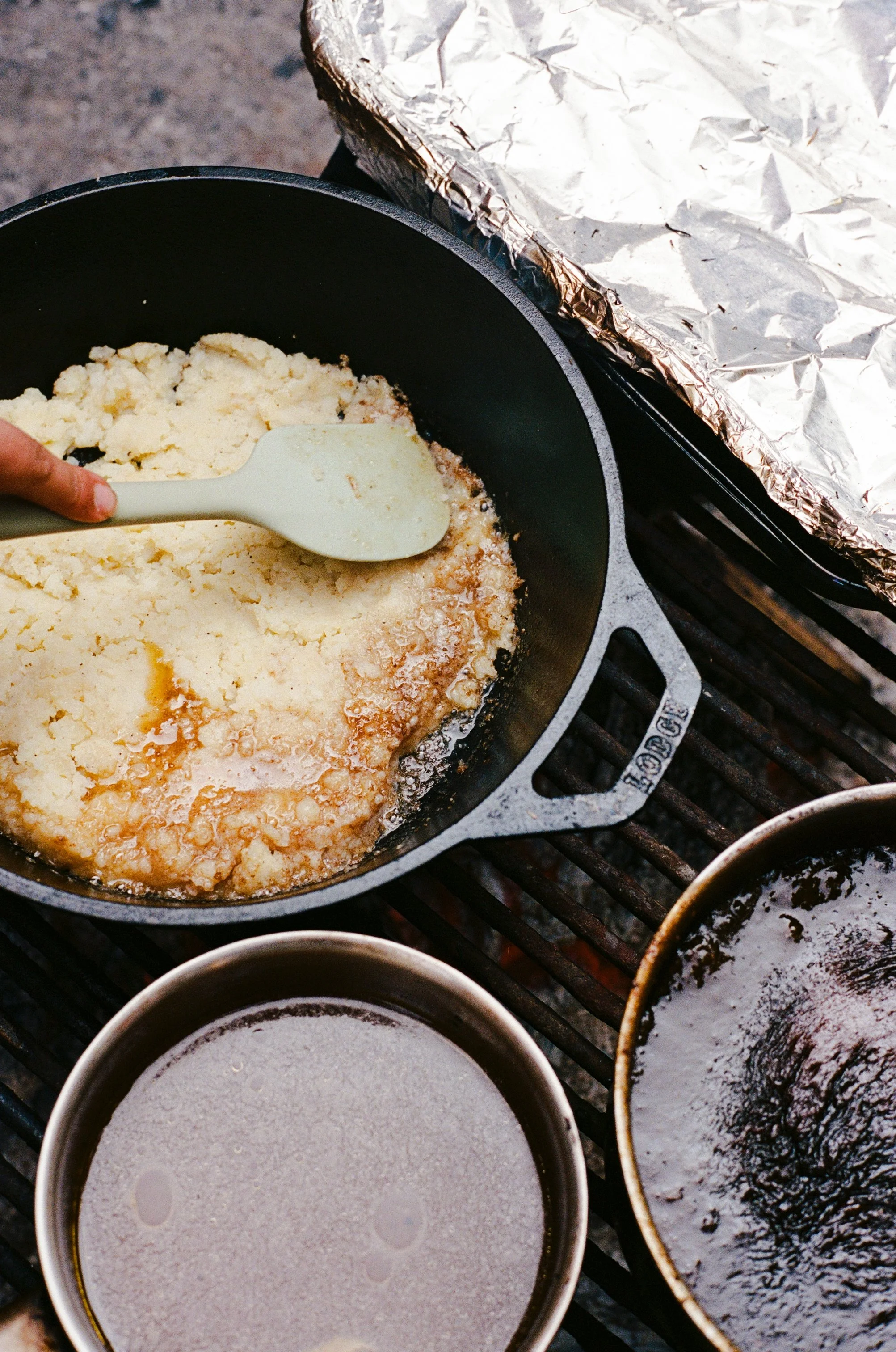 A person is cooking mashed potatoes in a black skillet on a grill, with aluminum foil covering part of the grill. There are two small pots nearby, one with a dark liquid and the other with a light liquid.