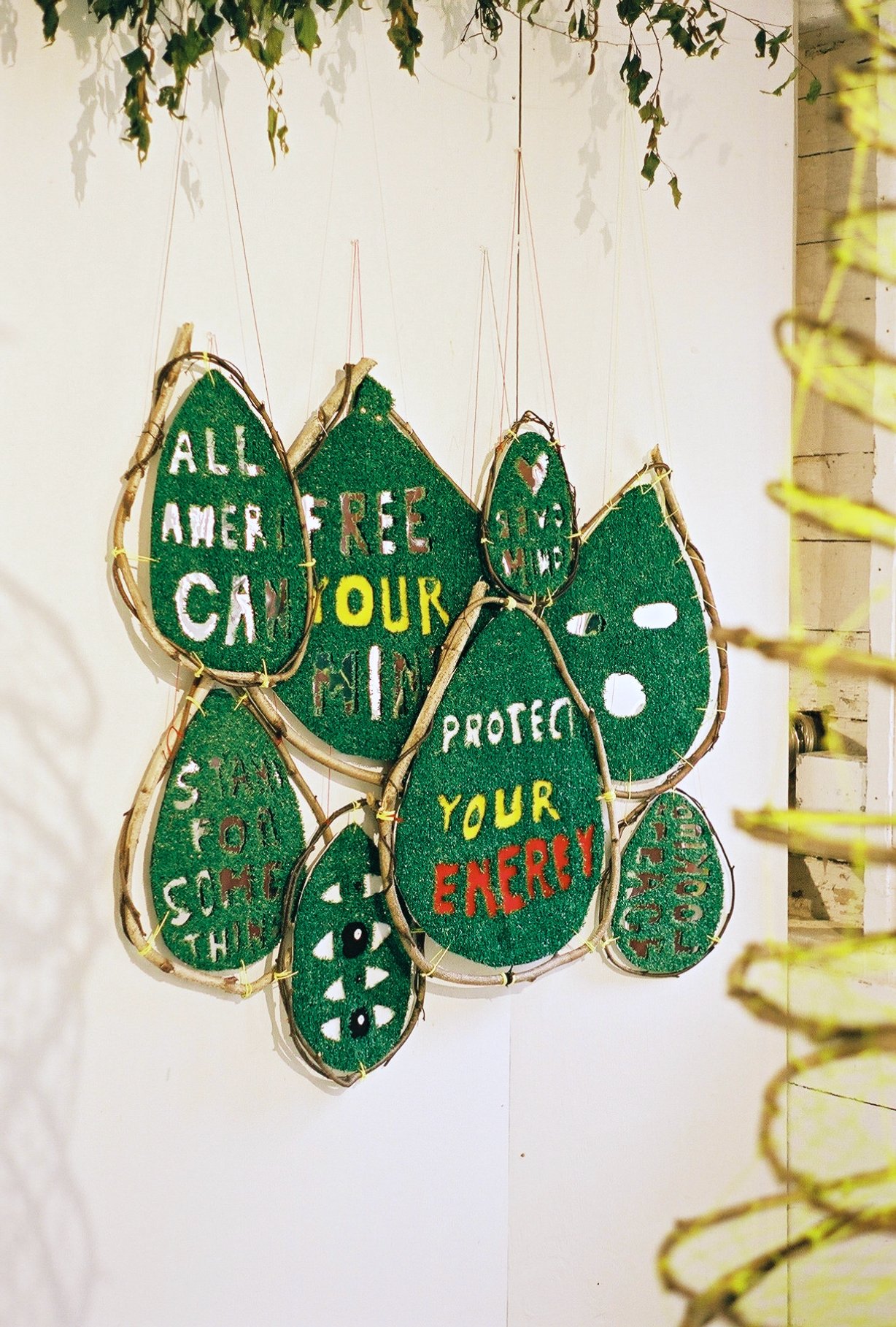 Decorative wall hanging with green leaf-shaped pieces displaying motivational messages in colorful letters, such as "ALL AMERICA CAN," "FREE," "PROTECT YOUR ENERGY," and others, attached to a wooden frame.