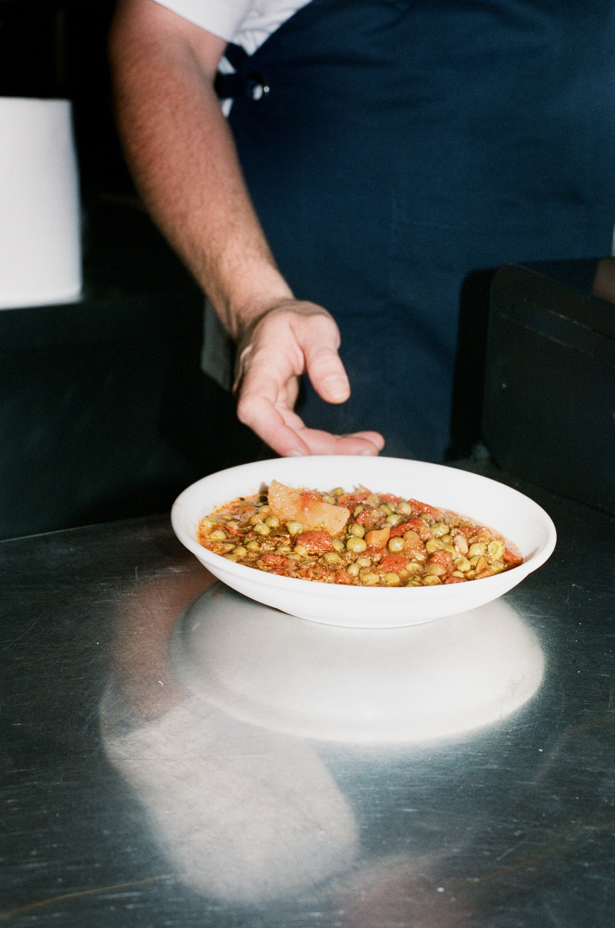A server presenting a bowl of vegetable soup with green peas, carrots, and potatoes on a stainless steel counter.