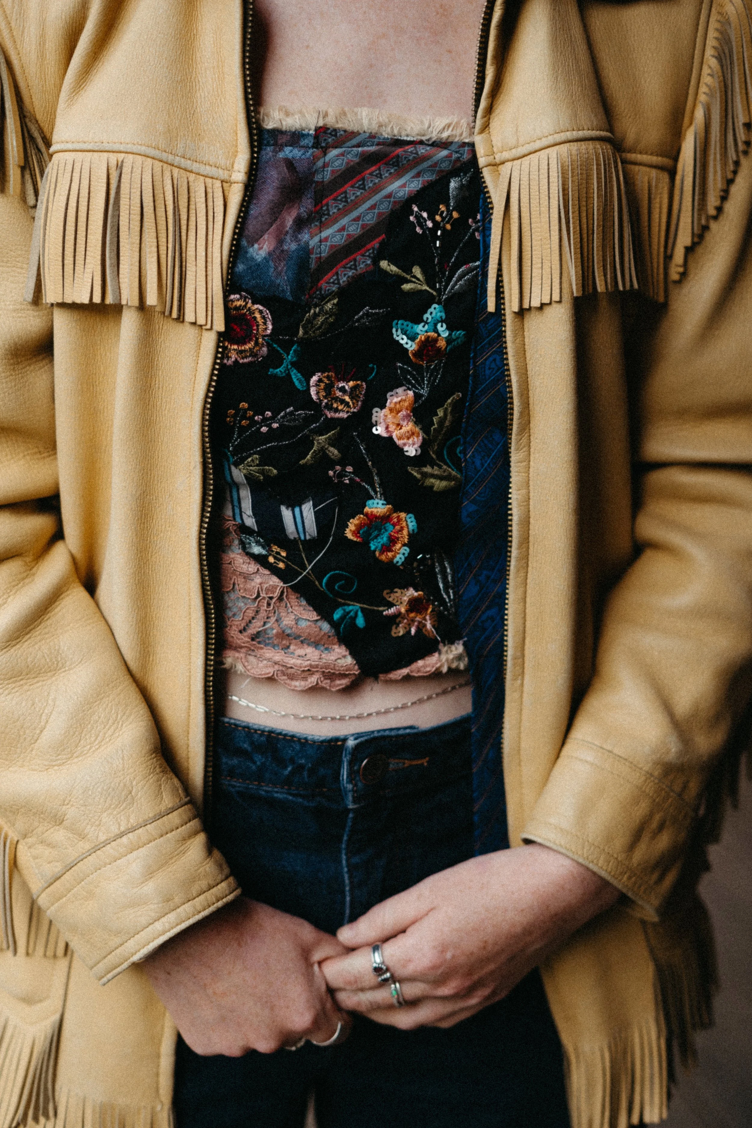 Close-up of a person's midsection wearing a tan fringed leather jacket, floral embroidery top, and dark jeans.