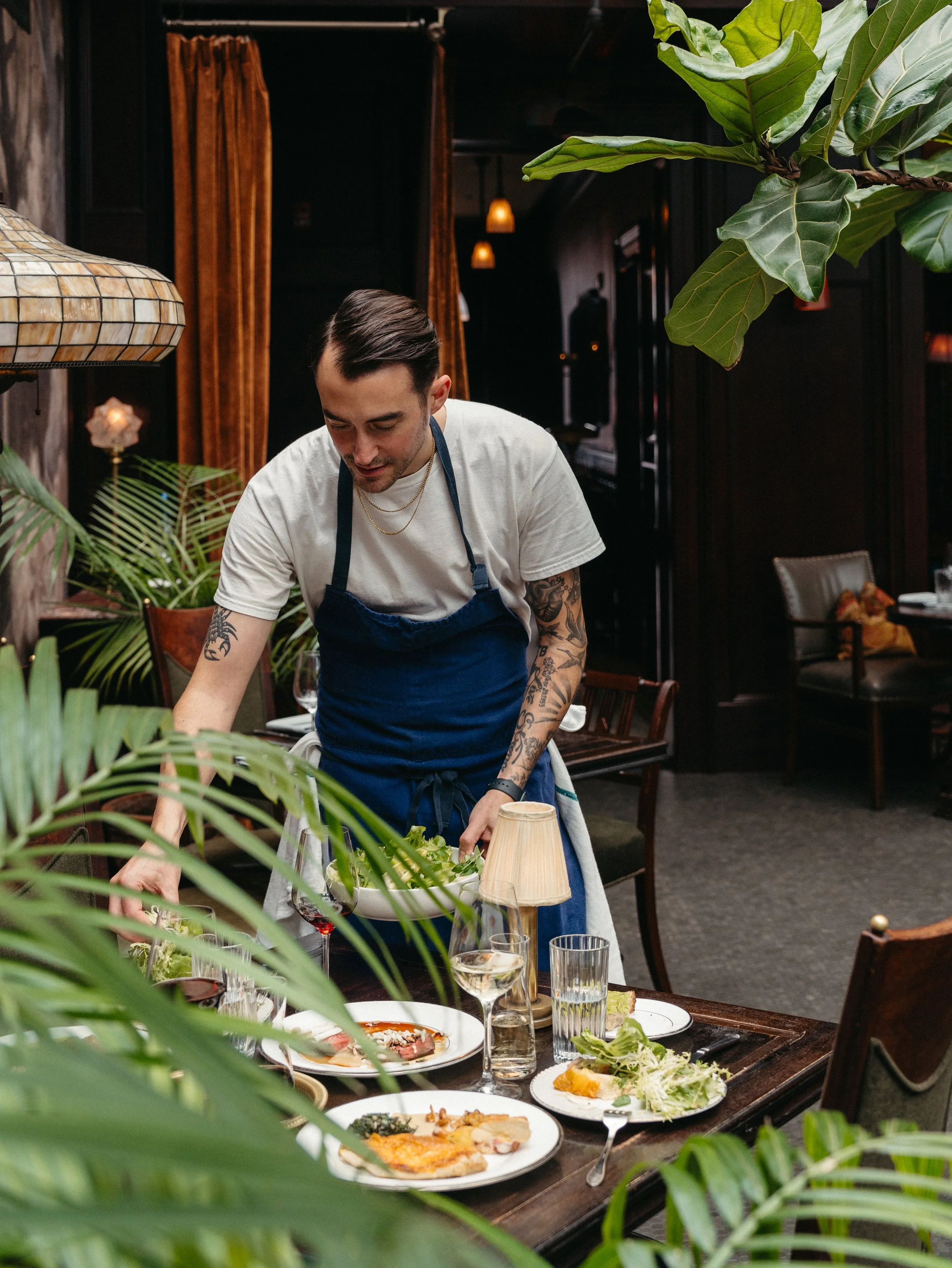 A male chef in a white T-shirt and blue apron preparing food at a table in a restaurant with dark walls, wooden furniture, and greenery.