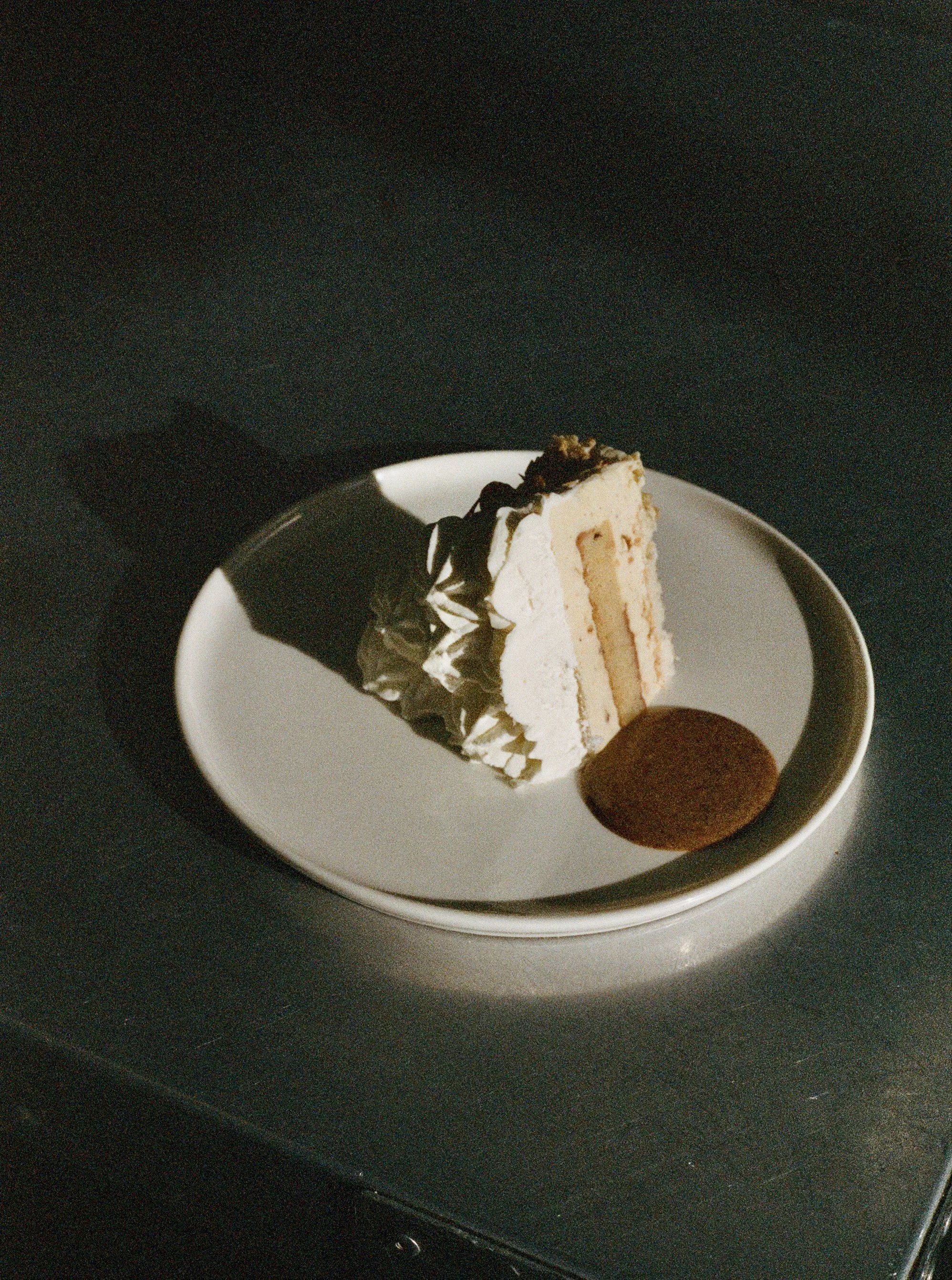 A slice of layered vanilla cake with white frosting and chocolate decorations on a white plate, accompanied by a sauce, against a dark background.