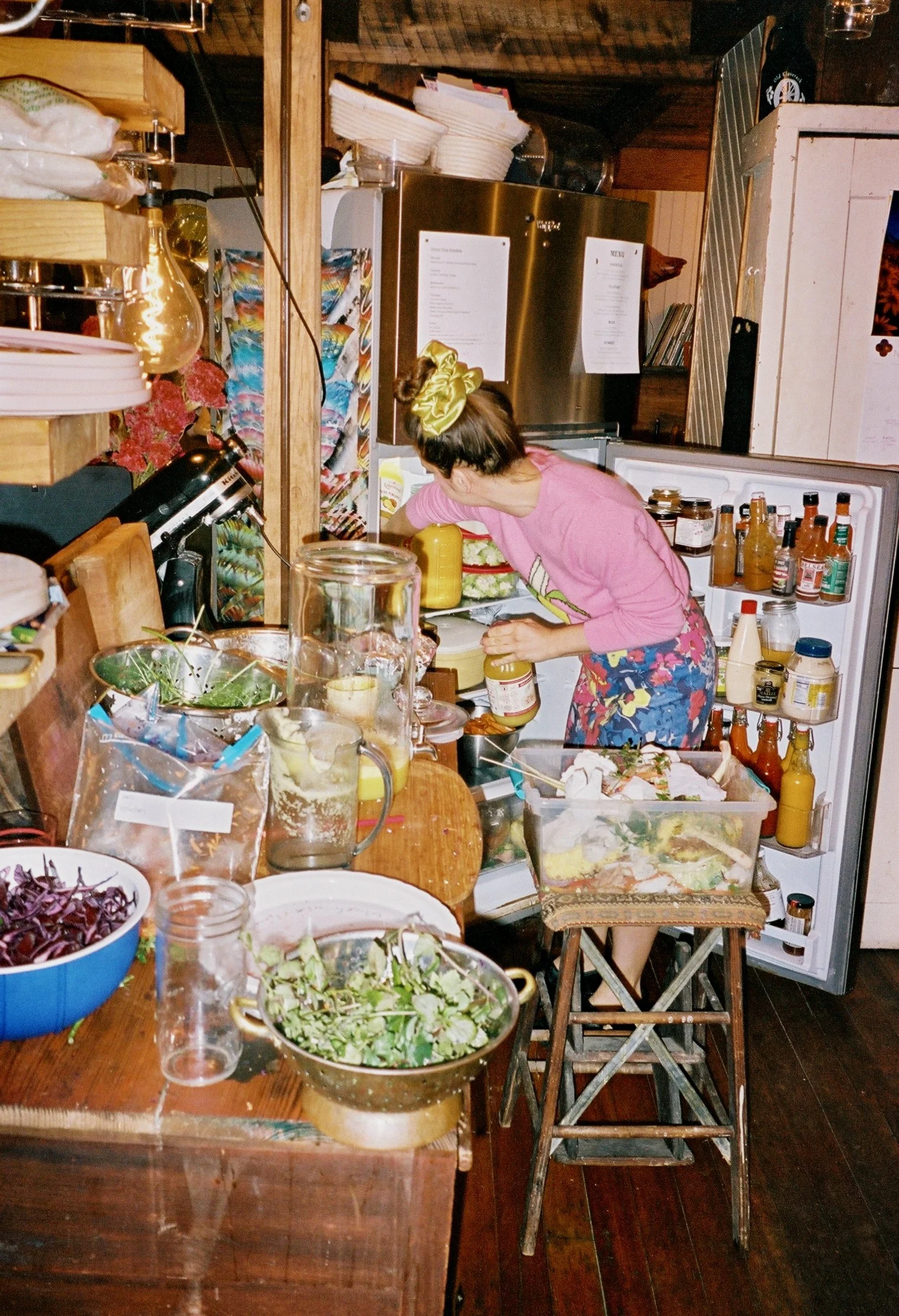 A woman in a pink shirt and floral skirt is organizing condiments inside an open refrigerator in a cozy kitchen with wood paneling and a busy wooden table filled with salads and kitchen items.