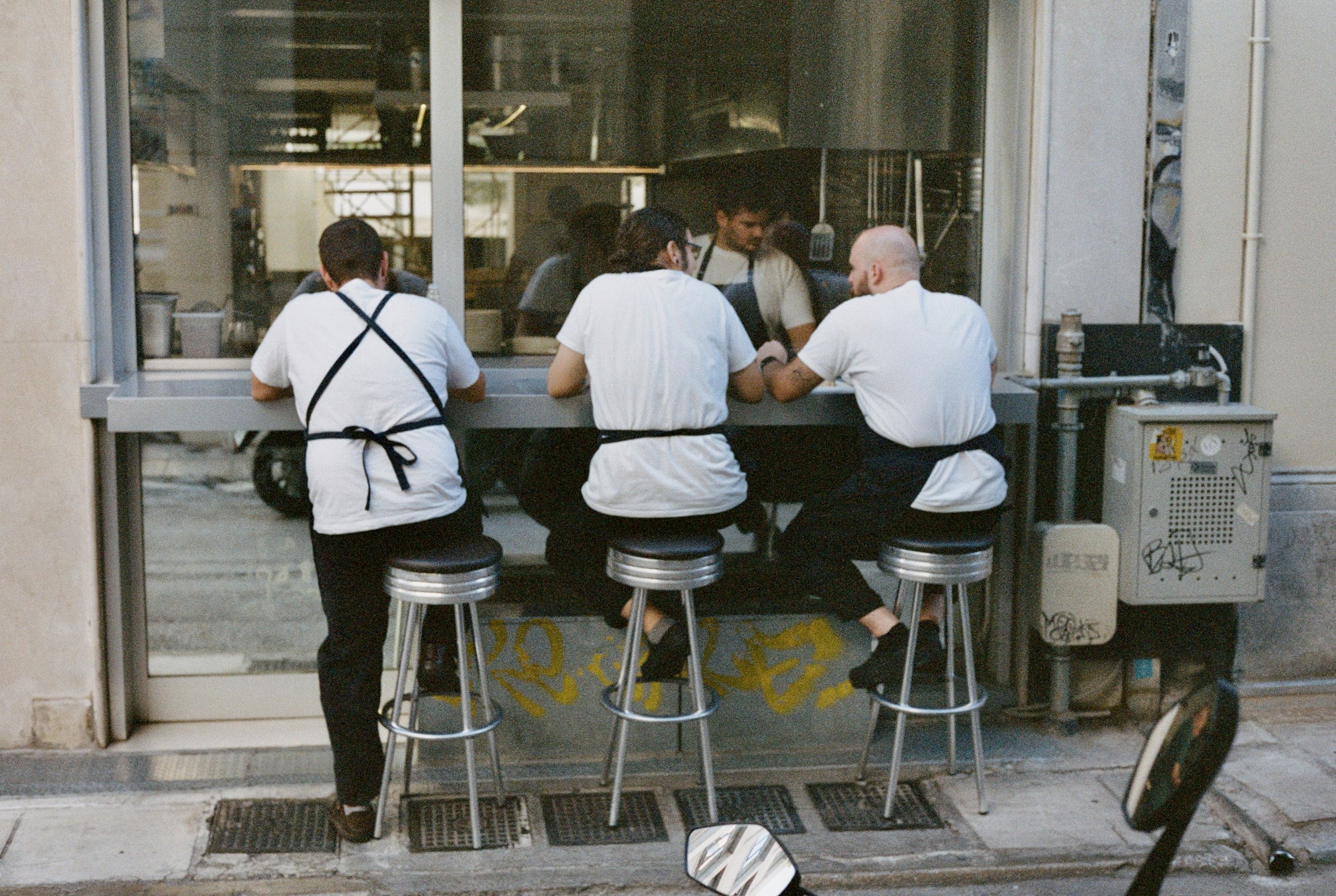Three people sitting at a counter window of a restaurant seen from outside. They are all wearing white shirts and black aprons, with one standing and two sitting on counter stools. Inside the kitchen, several staff are preparing food.