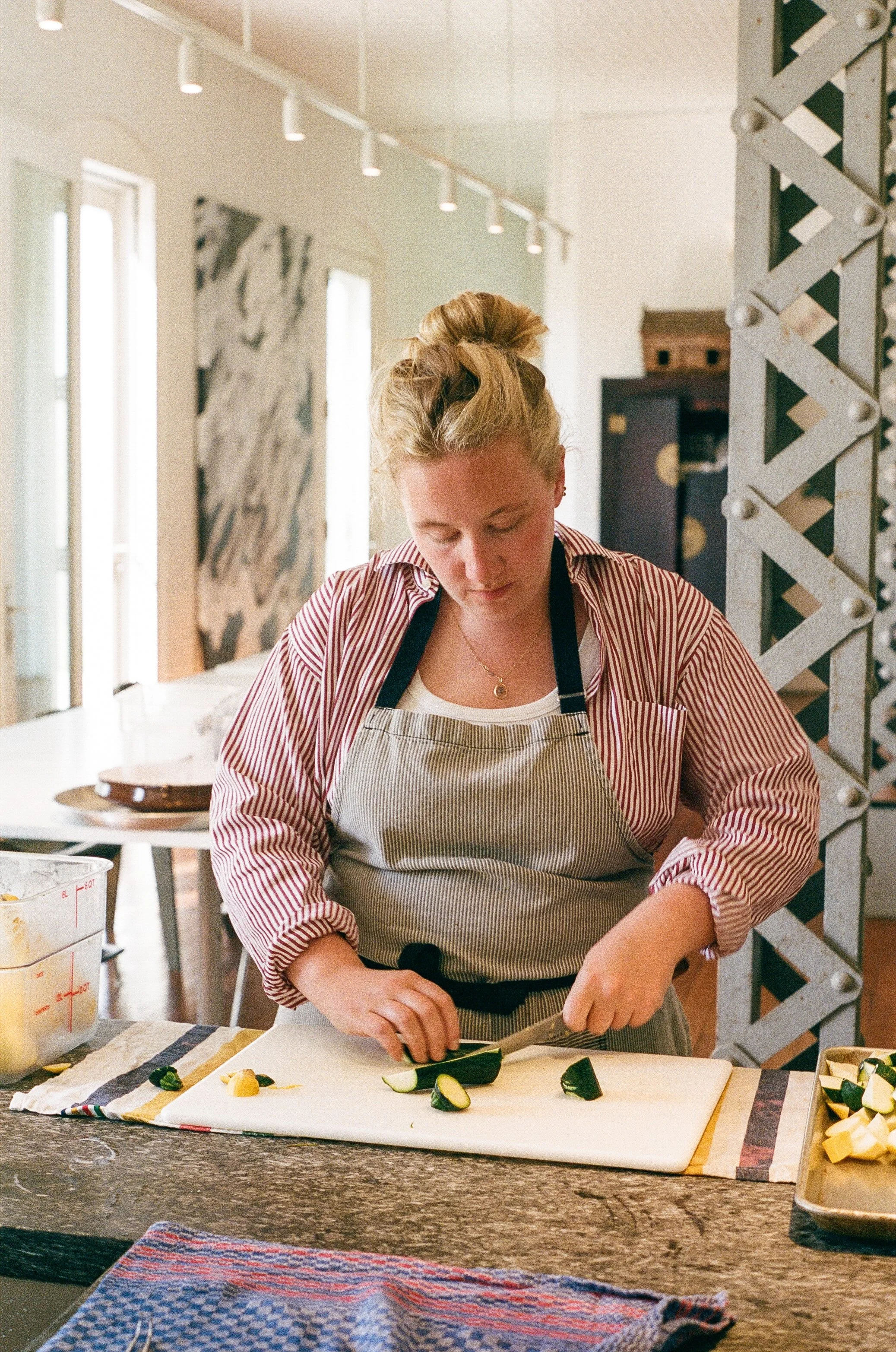 A woman with blonde hair tied up, wearing a striped shirt and apron, is chopping cucumbers on a white cutting board in a bright kitchen.